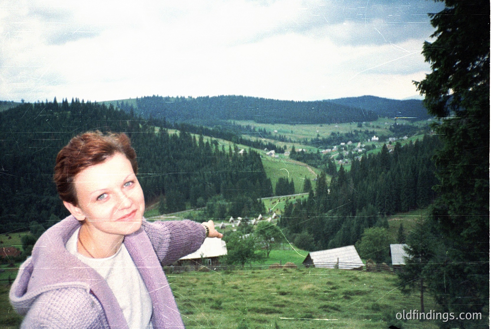 A woman poses in a lush, forested valley, pointing toward a distant village. She wears a purple sweater over a white top, set against dense coniferous trees and rolling green hills. Likely Eastern Europe, 1980s-1990s.