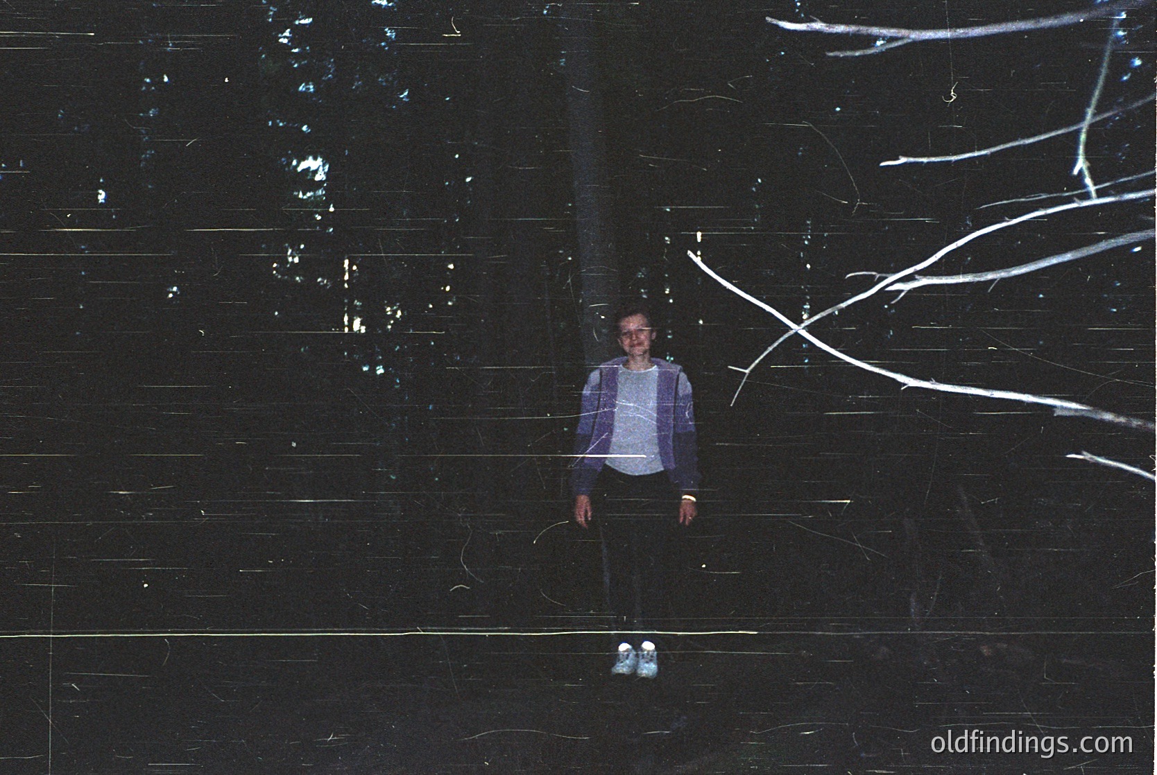 Portrait of a person standing under illuminated tree branches against a dark, textured wall. Light trails create dynamic patterns. Minimalist composition with monochromatic tones.