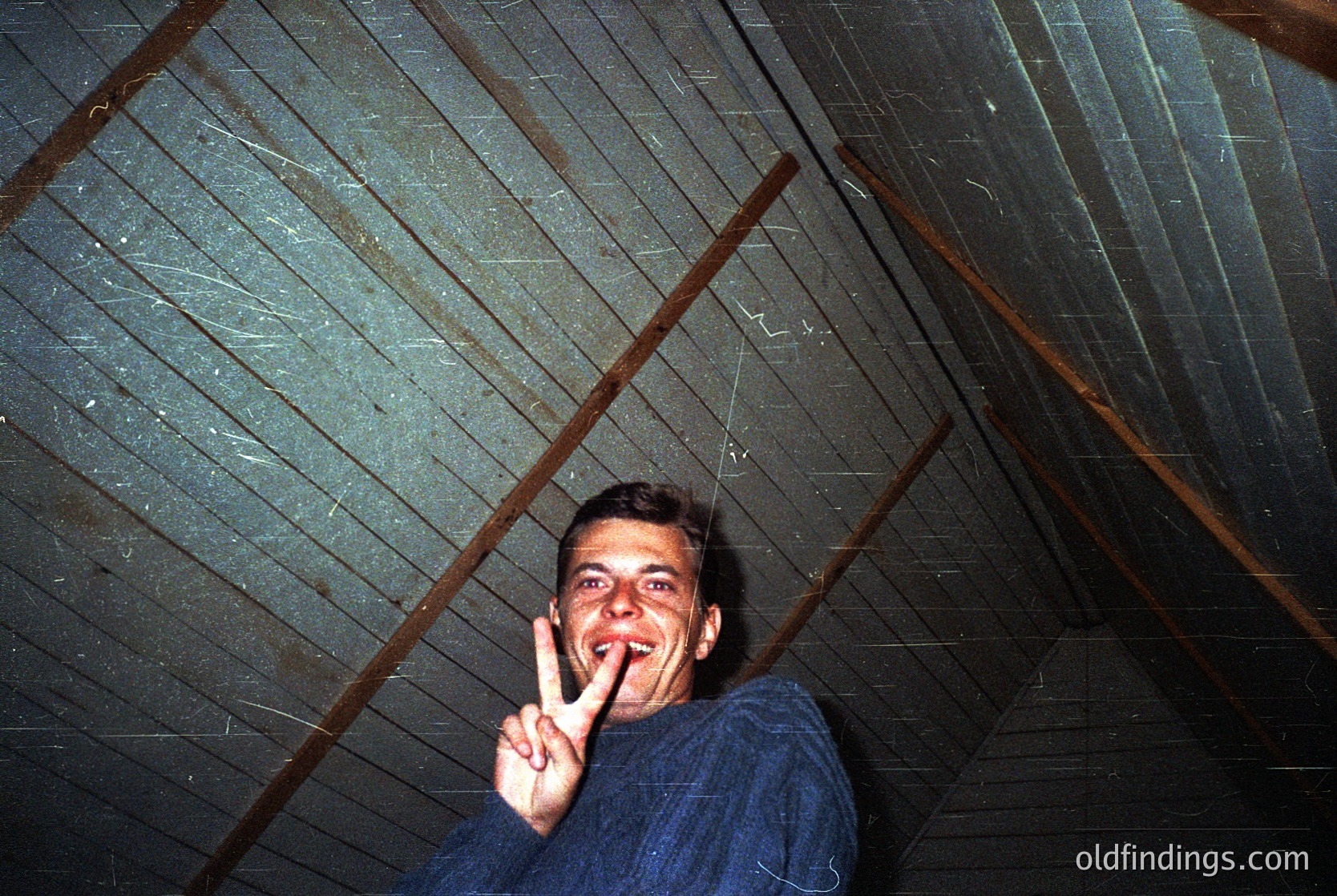 Vintage indoor shot of a man in a blue sweater making a peace sign under a rustic wooden ceiling with exposed beams. Likely 1970s–1980s, possibly a rural or industrial setting.