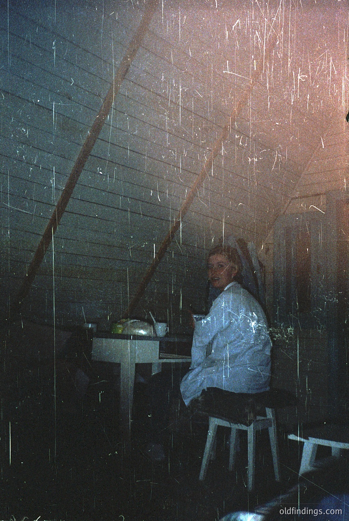 A person sits alone in a dimly lit, modern indoor space with sleek concrete walls and a minimalist stool. Rain-like light streams through transparent vertical panels, creating a dynamic visual effect. The scene evokes a futuristic café or gallery setting.