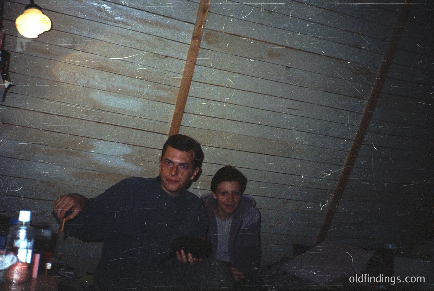 Vintage indoor shot of two men in a rustic wooden cabin, likely 1970s–1980s. Dark, textured planks and a single bulb illuminate the scene. One man holds a wooden spoon, the other rests on a wooden surface. Bottle and mug suggest a casual gathering.