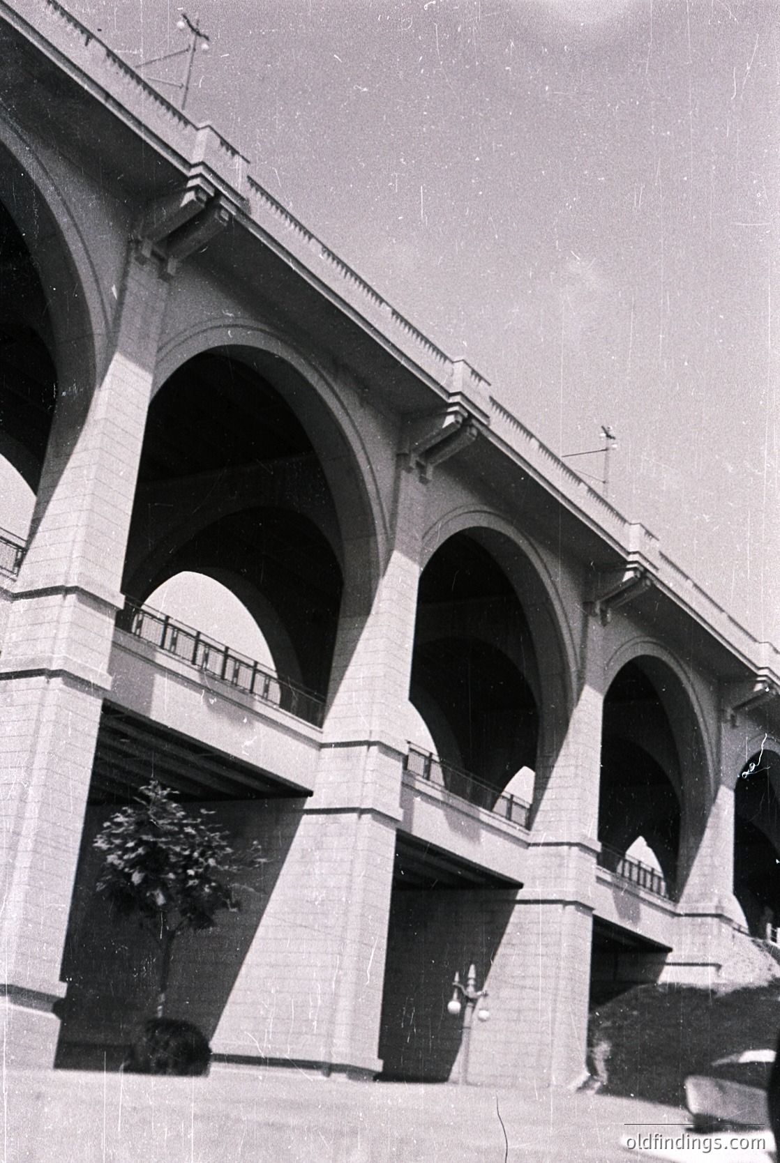 Neoclassical stone bridge with three prominent arches and balustraded upper level, likely mid-20th century. Architectural details include decorative corbels and a central ventilation cupola. Reflections in wet pavement suggest recent rain.
