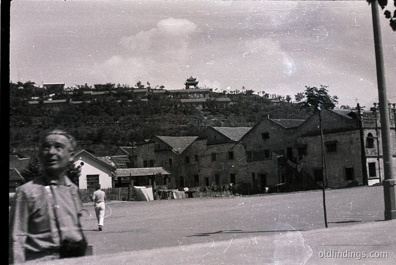 Vintage black-and-white street scene featuring traditional East Asian architecture with tiled roofs and wooden structures. Foreground shows blurred pedestrians; midground reveals a low fence and open courtyard. Background includes a hillside with dense foliage and a temple-like building atop. Likely *(Note: Specific location unverifiable without context, but architectural style suggests Hong Kong or similar region.)*