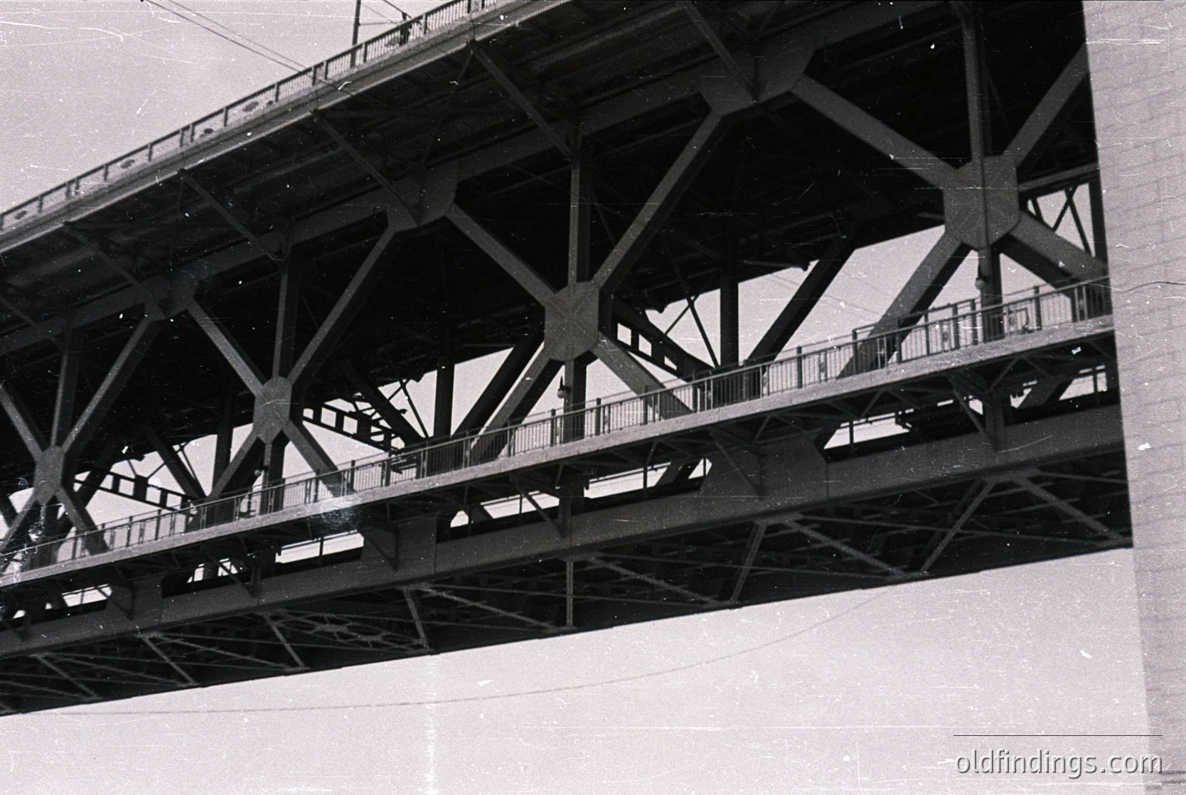 Black-and-white industrial bridge featuring riveted steel trusses and walkway railings, likely mid-20th century. Structural design showcases truss beams and diagonal supports, indicative of early-to-mid 1900s engineering. Potential use for transportation or utility infrastructure.