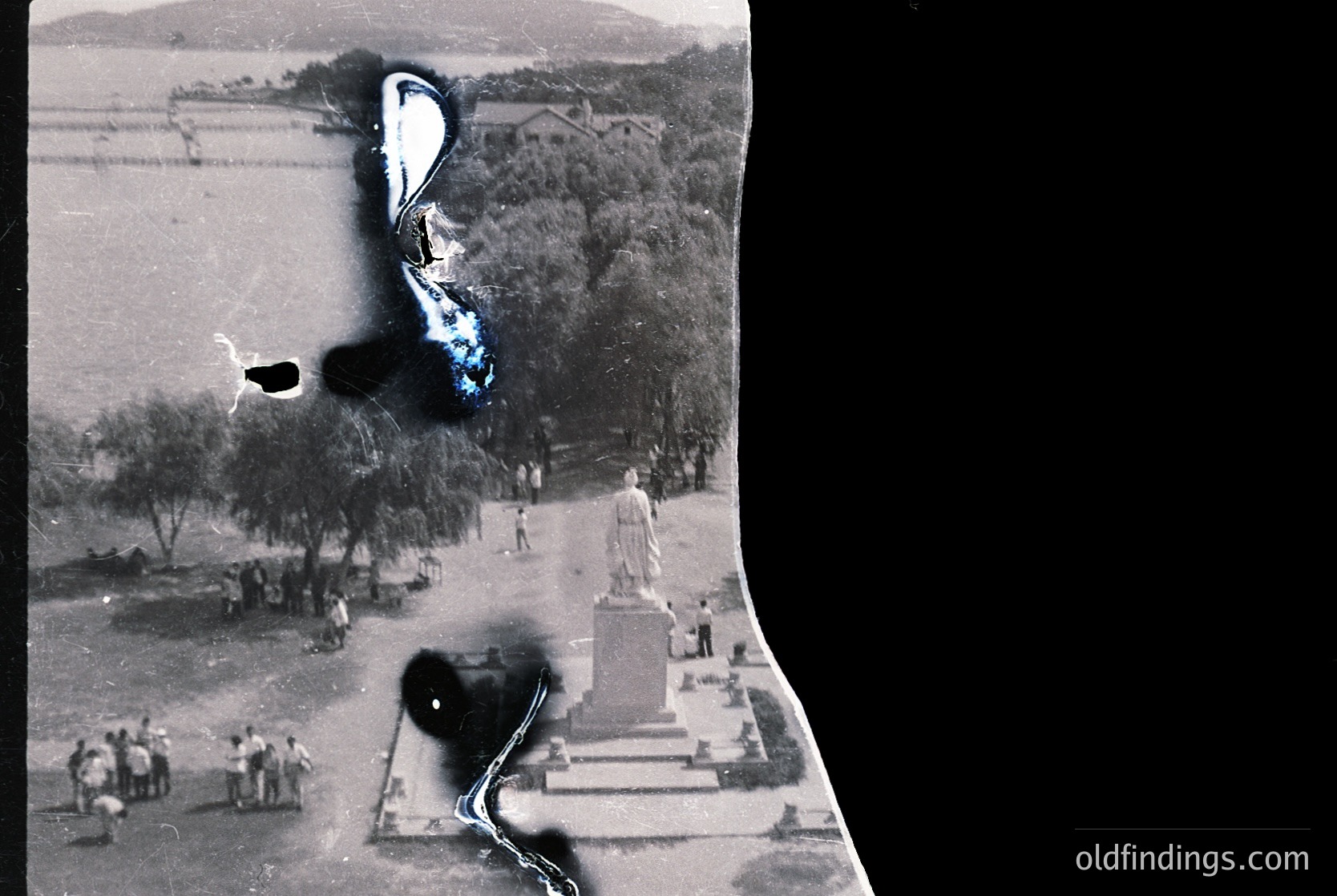 Vintage aerial photo of a public square with a prominent statue pedestal and figures in mid-20th century attire. Agricultural fields and tree-lined pathways visible. Damaged edges suggest aged film or scanning artifacts.