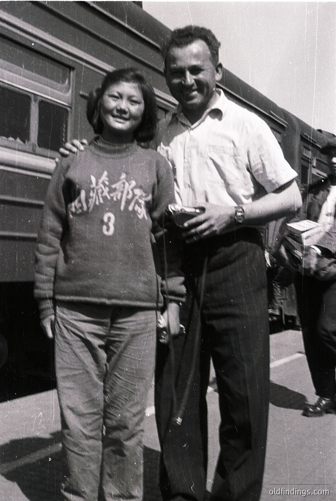 Black-and-white photo of two individuals posing near a vintage train car, likely mid-20th century. The woman wears a sweater with Japanese text and number "3," paired with wide-leg trousers. The man holds a small object in his hand and wears a button-up shirt with rolled sleeves. Background suggests a train station or transport hub.