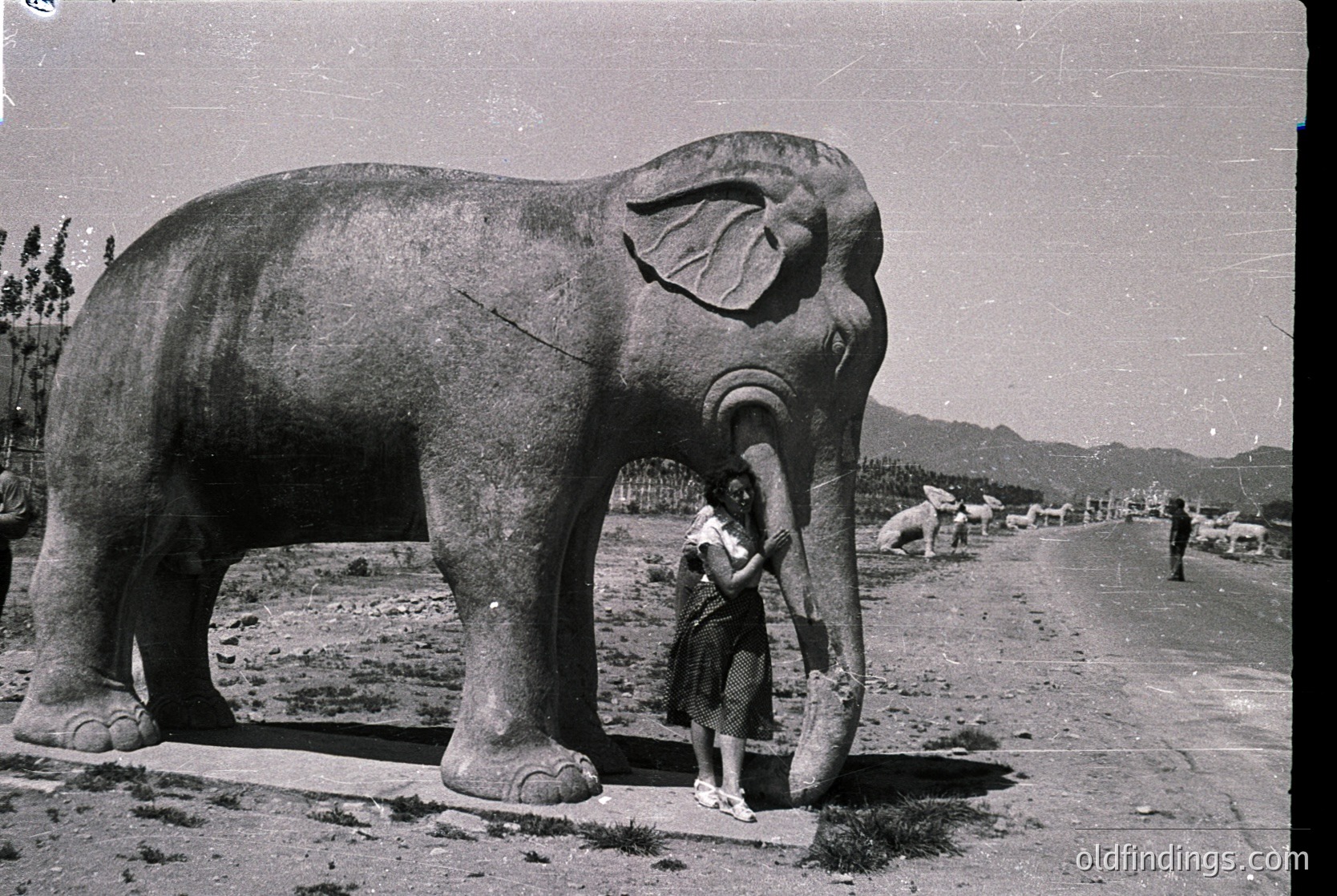 Vintage black-and-white photo of a large concrete elephant statue with a child interacting, likely mid-20th century. Mountainous background suggests a public park or tourist site.