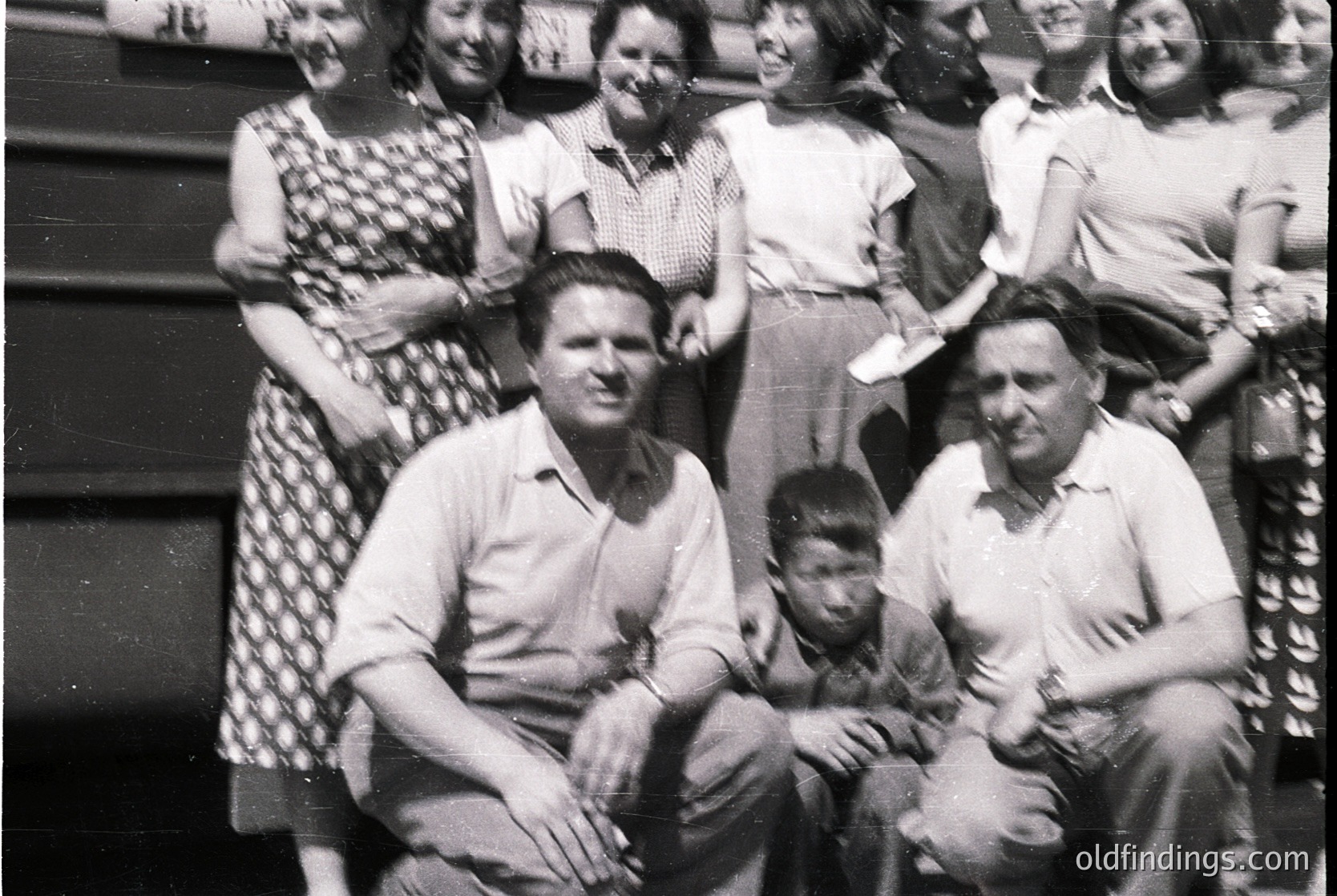 Black-and-white family portrait from the 1950s–1960s, featuring 9 individuals in formal attire. Men wear short-sleeve button-ups, women in patterned dresses and headscarves. Seated adults pose with a young boy in the center. Indoor setting with plain backdrop, likely a home or community hall.