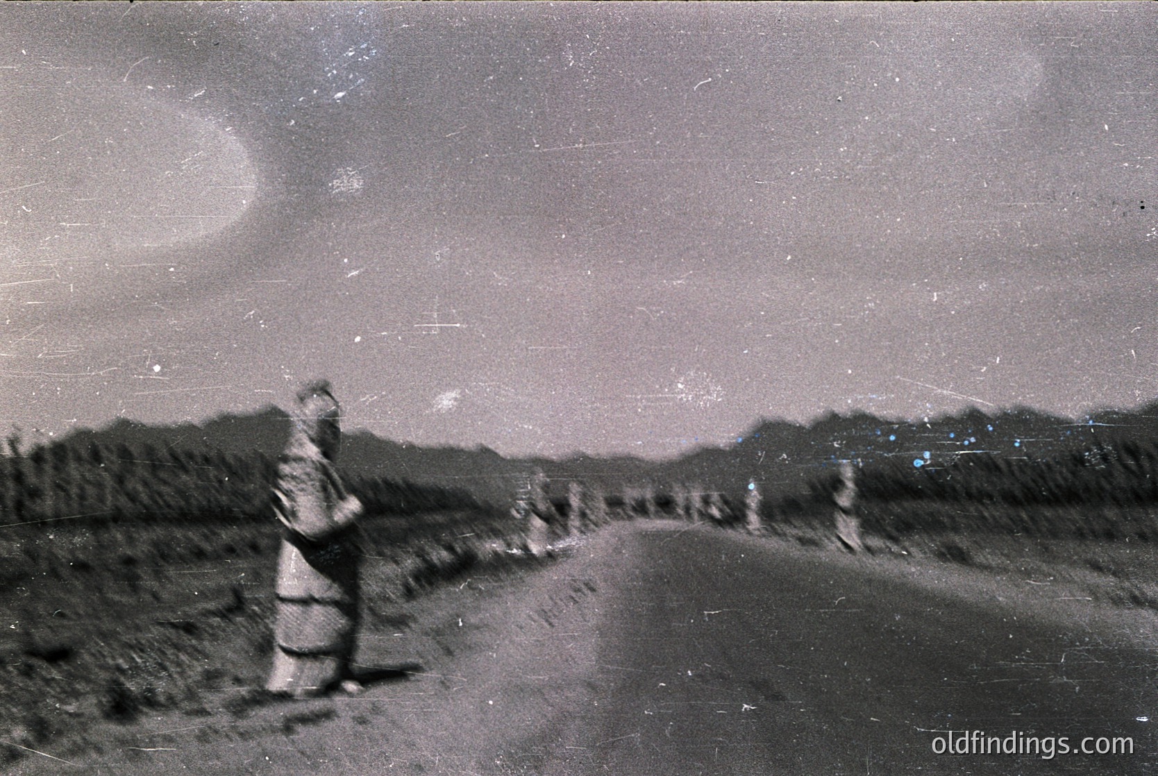 Vintage black-and-white photo of a lone figure in traditional alpine attire walking a narrow mountain road under a starry night sky. The silhouette suggests a high-altitude setting, likely the European Alps. The grainy texture and lighting indicate early 20th-century photography.