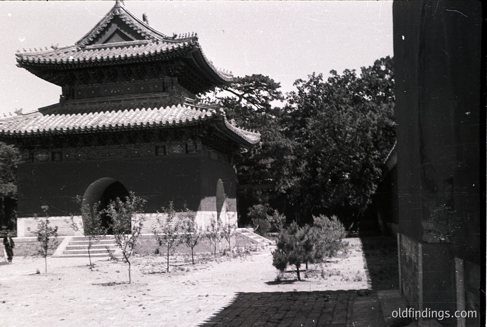 Traditional East Asian pavilion with upturned eaves and tiled roof, set on stone steps. Courtyard features young trees and minimal landscaping. Likely 20th-century architectural style, possibly or . Ideal for historical research or cultural design references.