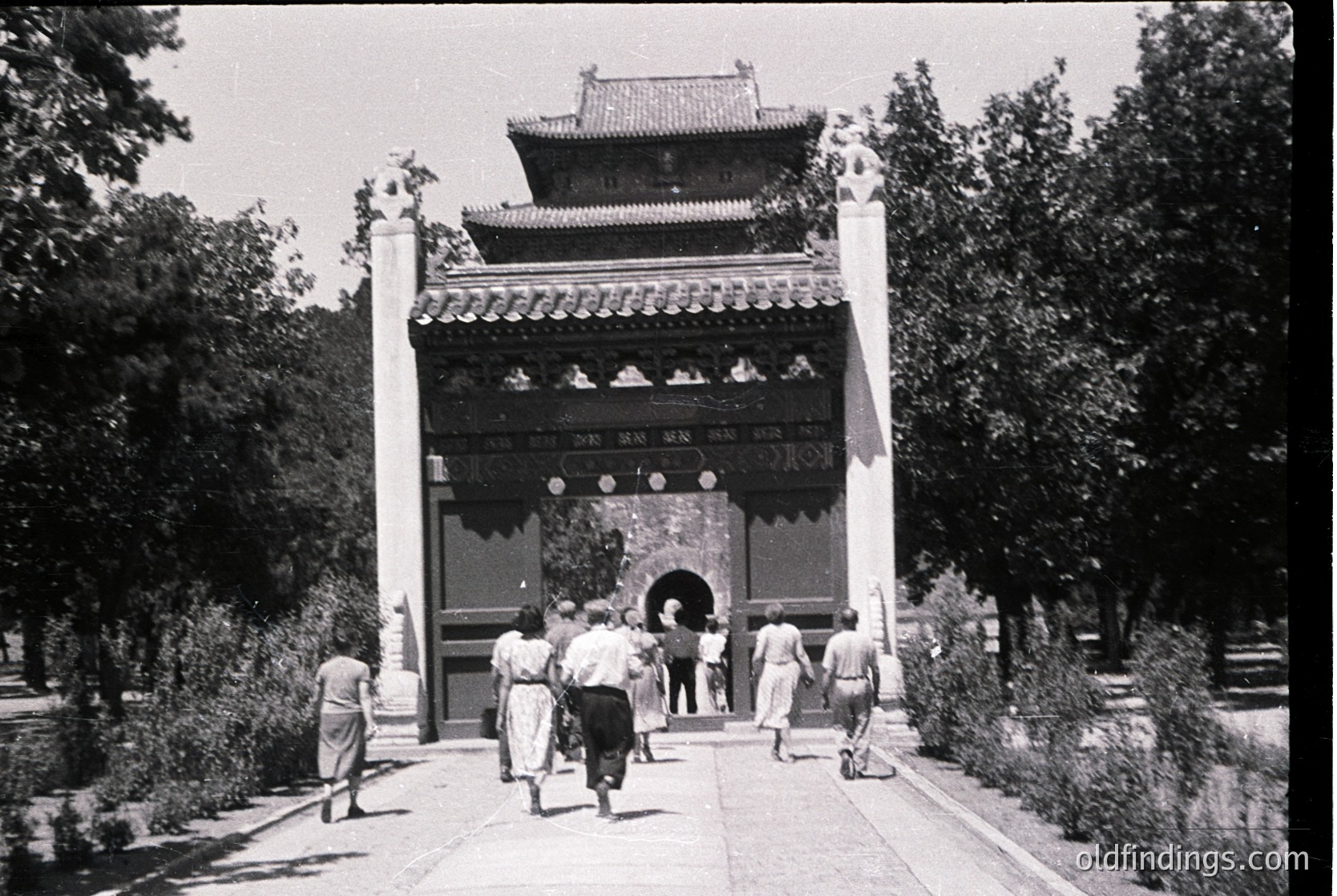 Black-and-white photograph of a traditional East Asian gate entrance with curved, tiled roof and Chinese characters above the archway. Six people in 1950s-era clothing (light jackets, trousers) walk toward the entrance through a landscaped pathway flanked by trees and shrubs.