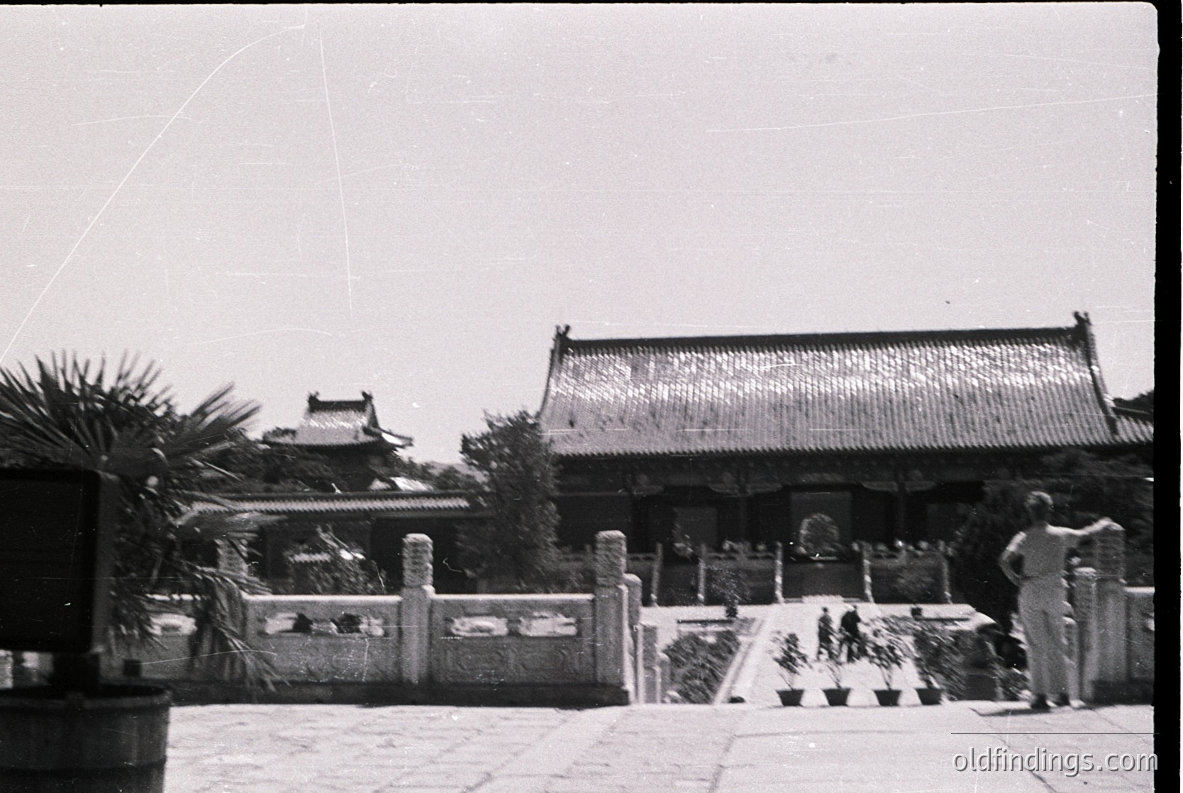 Black-and-white photograph of traditional East Asian temple courtyard, featuring tiled roofs, stone pathways, and potted plants. Architectural details include ornate eaves and a central gate. Likely mid-20th century, possibly or .