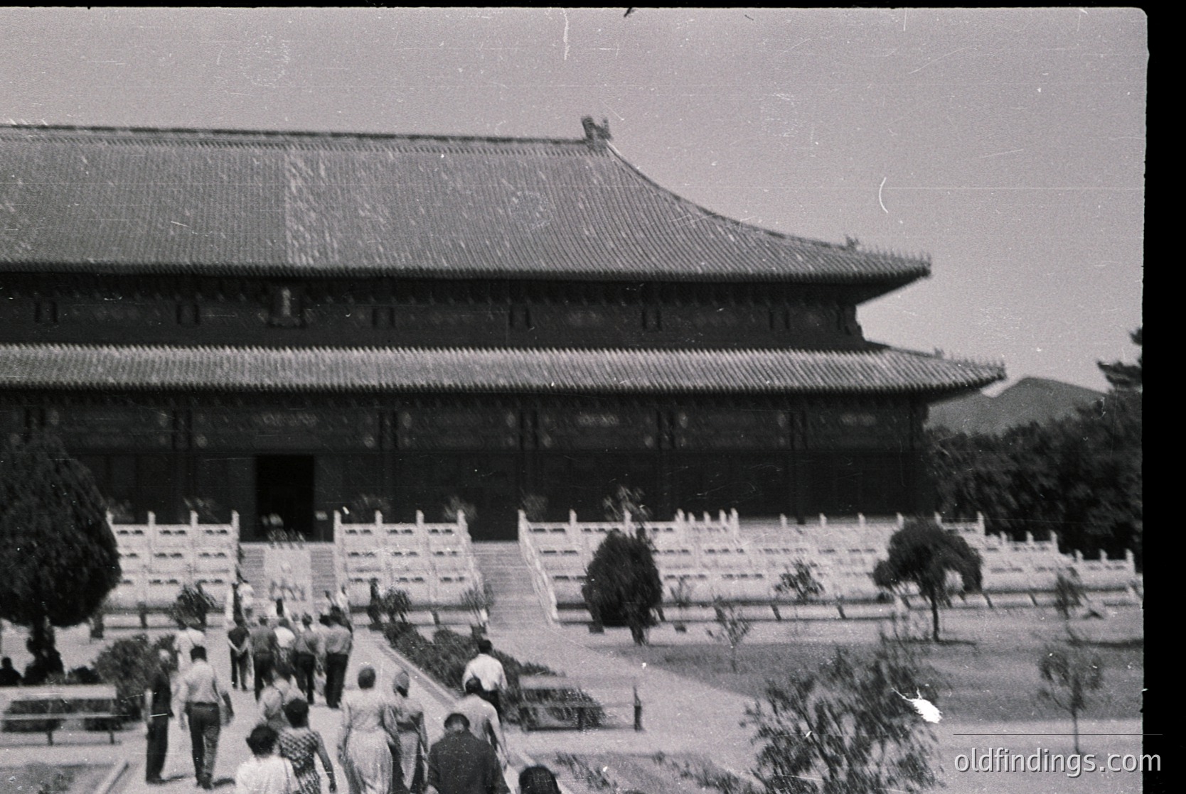 Vintage black-and-white photo of traditional East Asian palace architecture with curved, tiled roofs and white stone pathways. Symmetrical courtyard design with low white fences and sparse trees. Mid-20th century attire suggests or . Likely or due to architectural style.