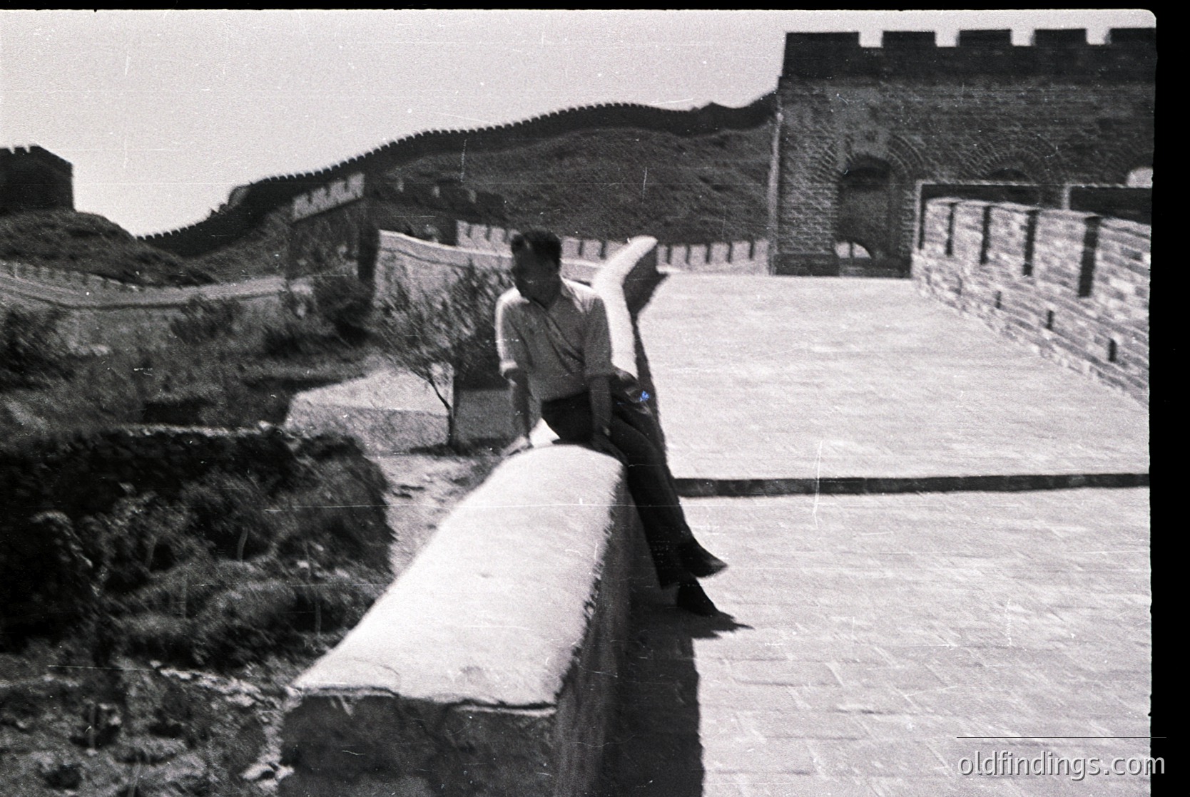 Black-and-white shot of a lone figure perched on a stone barrier atop the Great Wall of China, mid-gesture. Stone watchtowers and battlements stretch into the distance under clear skies.