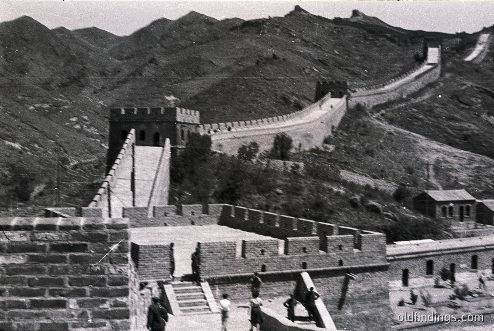 Historic black-and-white photo of the Great Wall of China, showcasing its iconic stone construction and watchtowers. The winding path ascends through rugged mountainous terrain, with a lone figure descending a staircase. Mid-20th century architectural heritage, likely