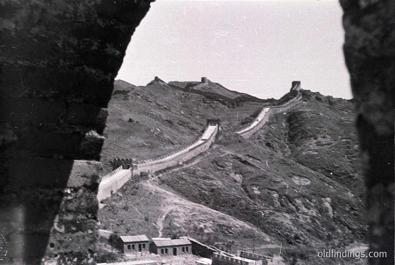 Black-and-white shot of the Great Wall of China winding through rugged terrain, framed by a stone arch. Mid-20th century construction style visible in foreground building.