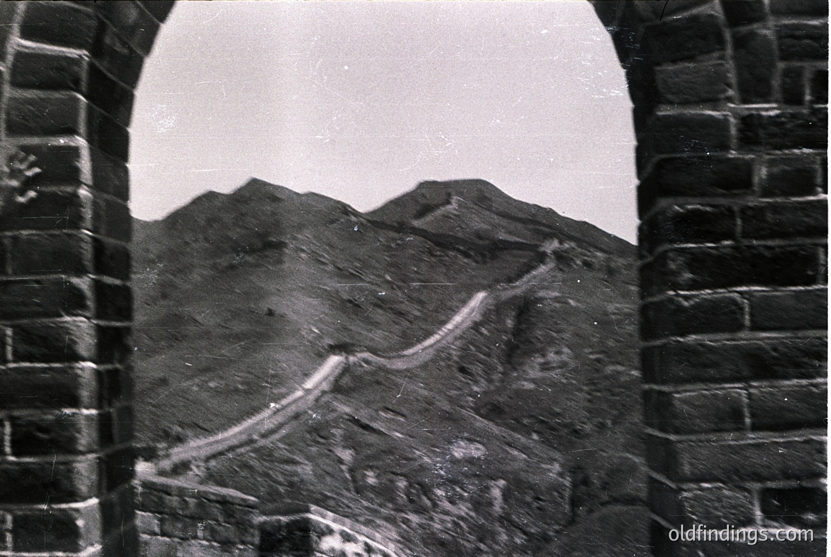 Black-and-white archival shot of the Great Wall winding through rugged mountains, framed by a stone archway. Distinctive brickwork and winding path highlight engineering of Ming/Qing dynasties. Likely 19th–early 20th century.