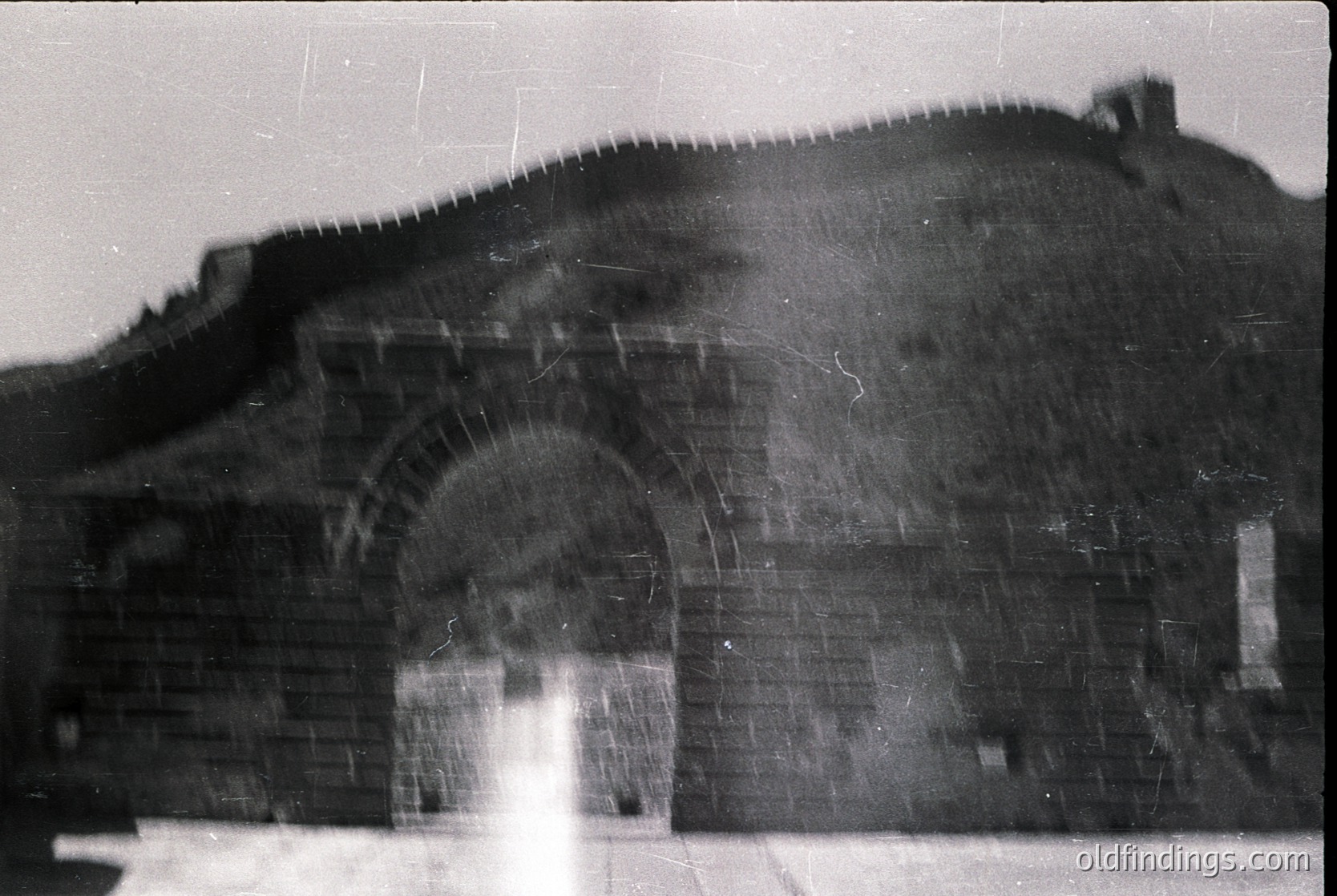 Vintage black-and-white photo of a large, arched brick gateway with weathered stonework. The archway appears to be part of a fortified structure, possibly a historic or industrial site. Overgrown vegetation and a sloped hilltop in the background suggest abandonment. Likely Eastern European, mid-20th century.