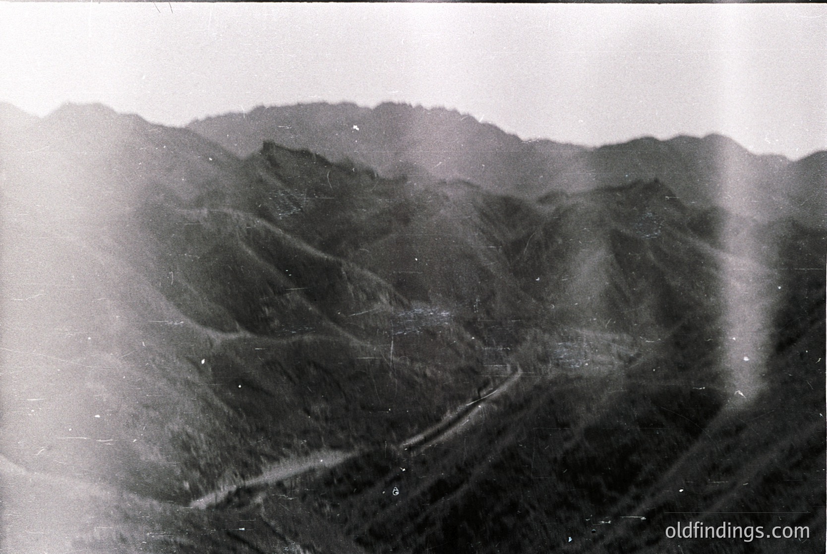 Vintage black-and-white aerial view of rugged mountain terrain with winding road cutting through steep slopes. Dense forest covers lower elevations, fading into rocky ridges. Likely mid-20th century due to grainy film texture.