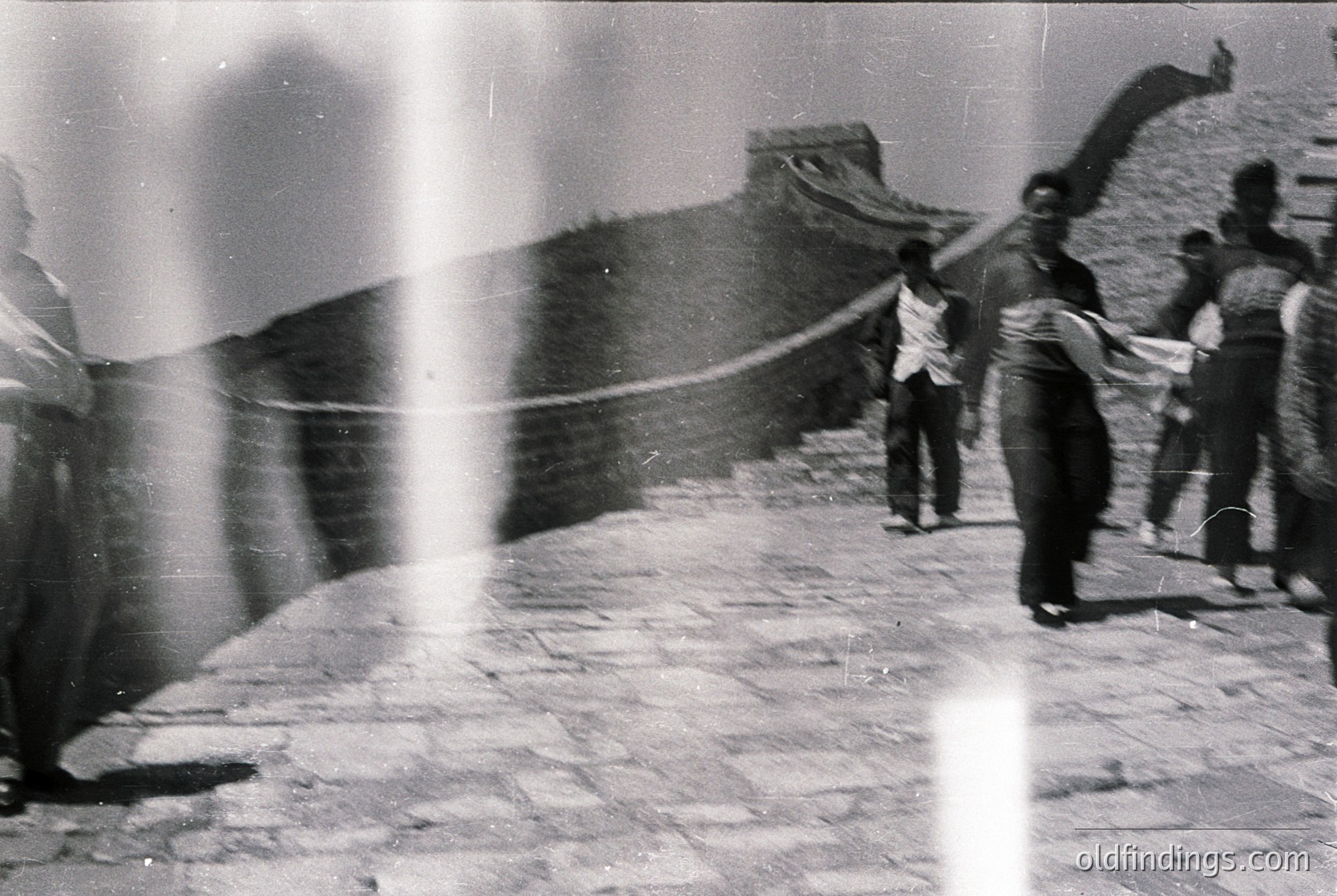 Blurred vintage black-and-white shot of the Great Wall of China with three figures in motion, likely mid-20th century. Stone pathway and watchtower ruins visible. Dynamic composition captures movement and historical architecture.