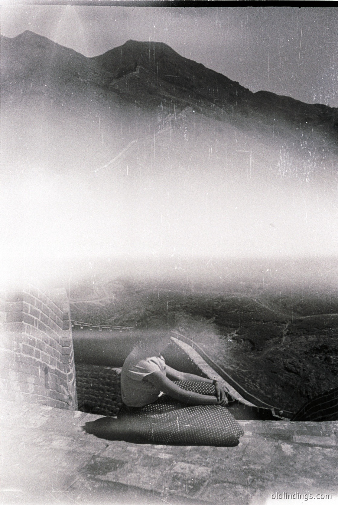 Vintage black-and-white photo of a lone figure seated on a stone ledge overlooking misty alpine peaks. The person wears patterned trousers and a loose top, suggesting mid-20th-century fashion. The rugged stone wall and misty landscape evoke a serene, contemplative mood.