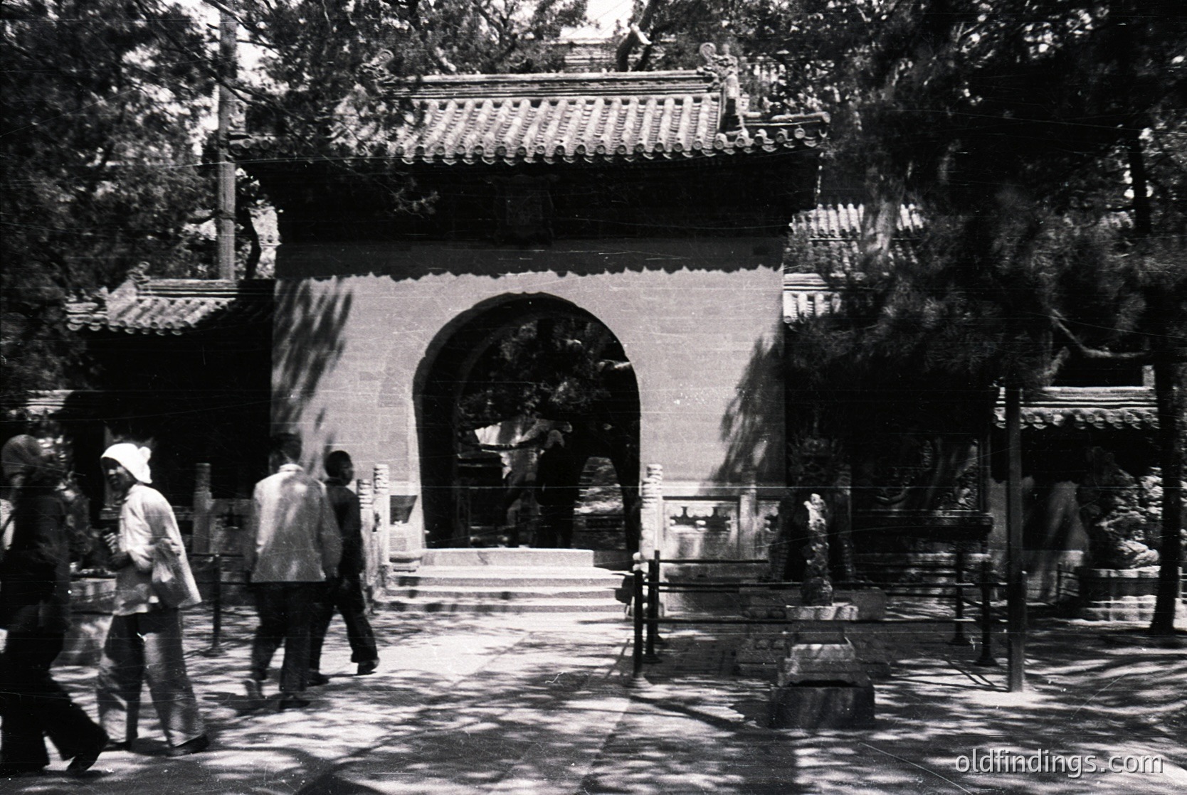 Traditional Chinese temple entrance with curved, tiled roof and stone archway. Visitors in 1970s-era clothing walk through shaded courtyard. Ornate lanterns and stone railings frame the pathway.