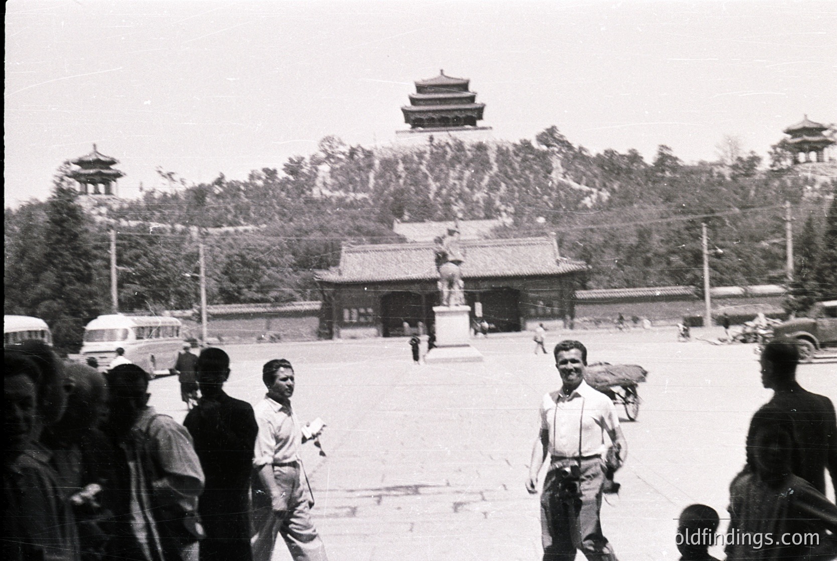 Classic black-and-white shot of Tiananmen Square, Beijing, featuring the iconic **Tiananmen Gate** in the background. Mid-20th century (1950s–1960s) attire and Soviet-style buses suggest post-liberation era. Crowd of pedestrians, including a man carrying a bundle, adds to the historical atmosphere.