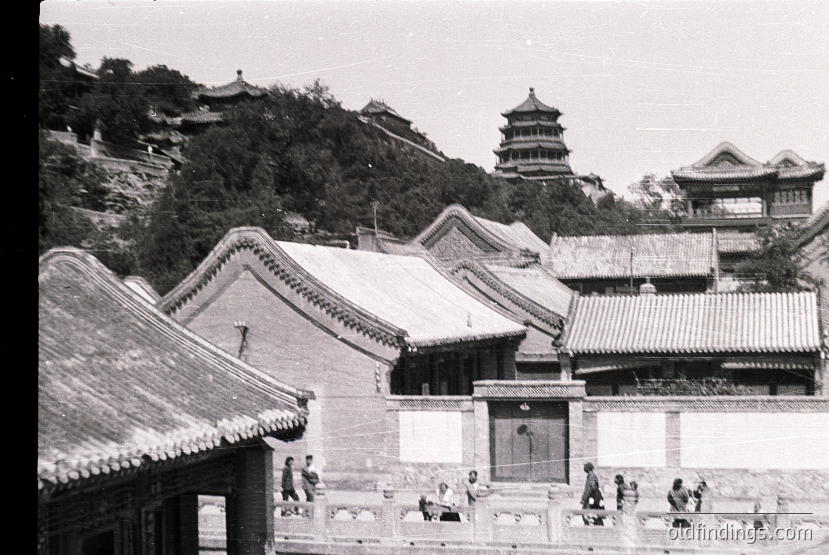 Classic Ming/Qing dynasty courtyard architecture with upturned eaves, set against a forested hillside. Mid-20th century black-and-white photo captures traditional Chinese pavilion and seated visitors, likely Beijing’s Summer Palace.