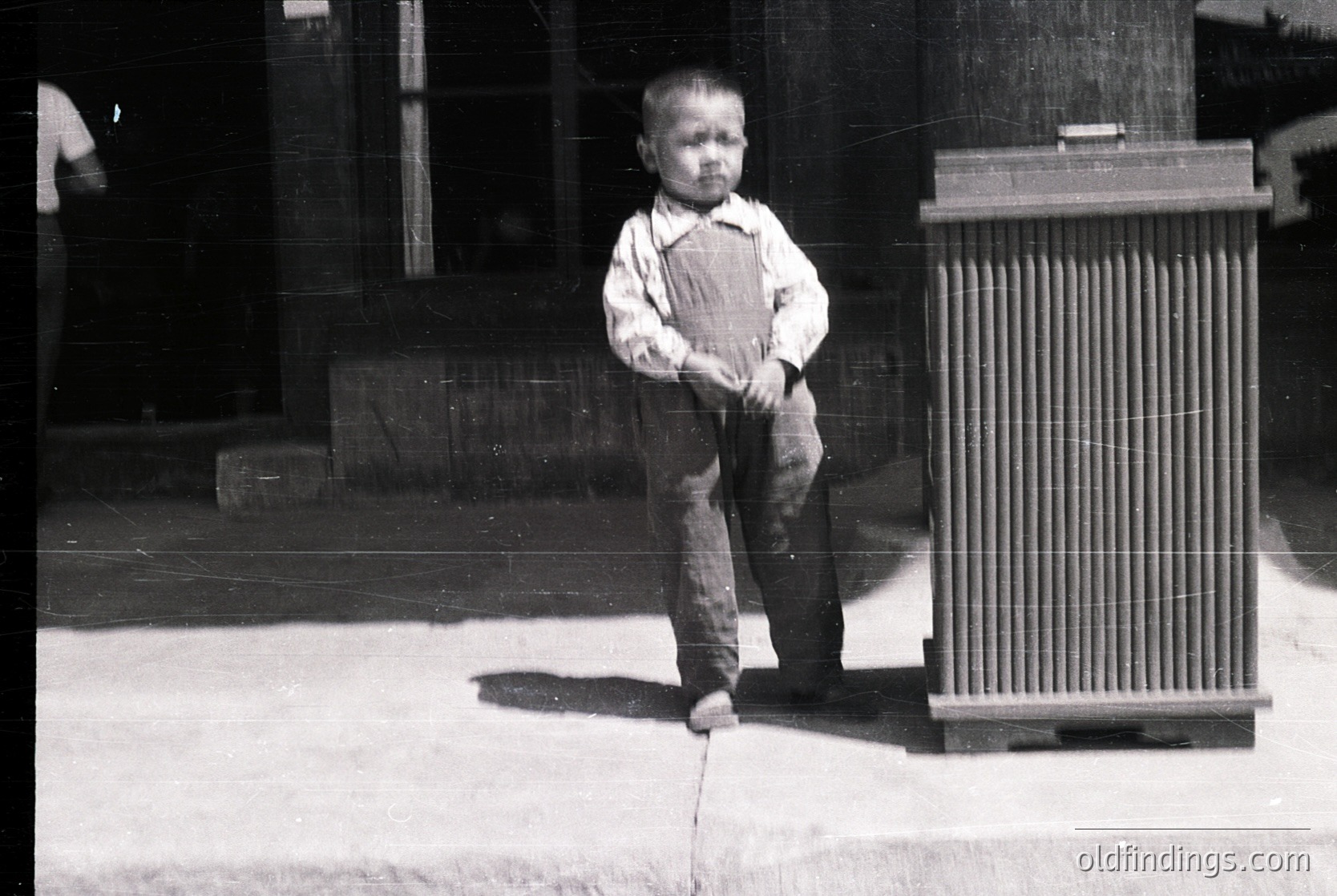 Young boy in early 20th-century attire—overalls, short-sleeved shirt, and sandals—standing on a concrete surface beside a vertical wooden slat fence. Blurry background suggests an urban or semi-urban setting, likely early 1900s.