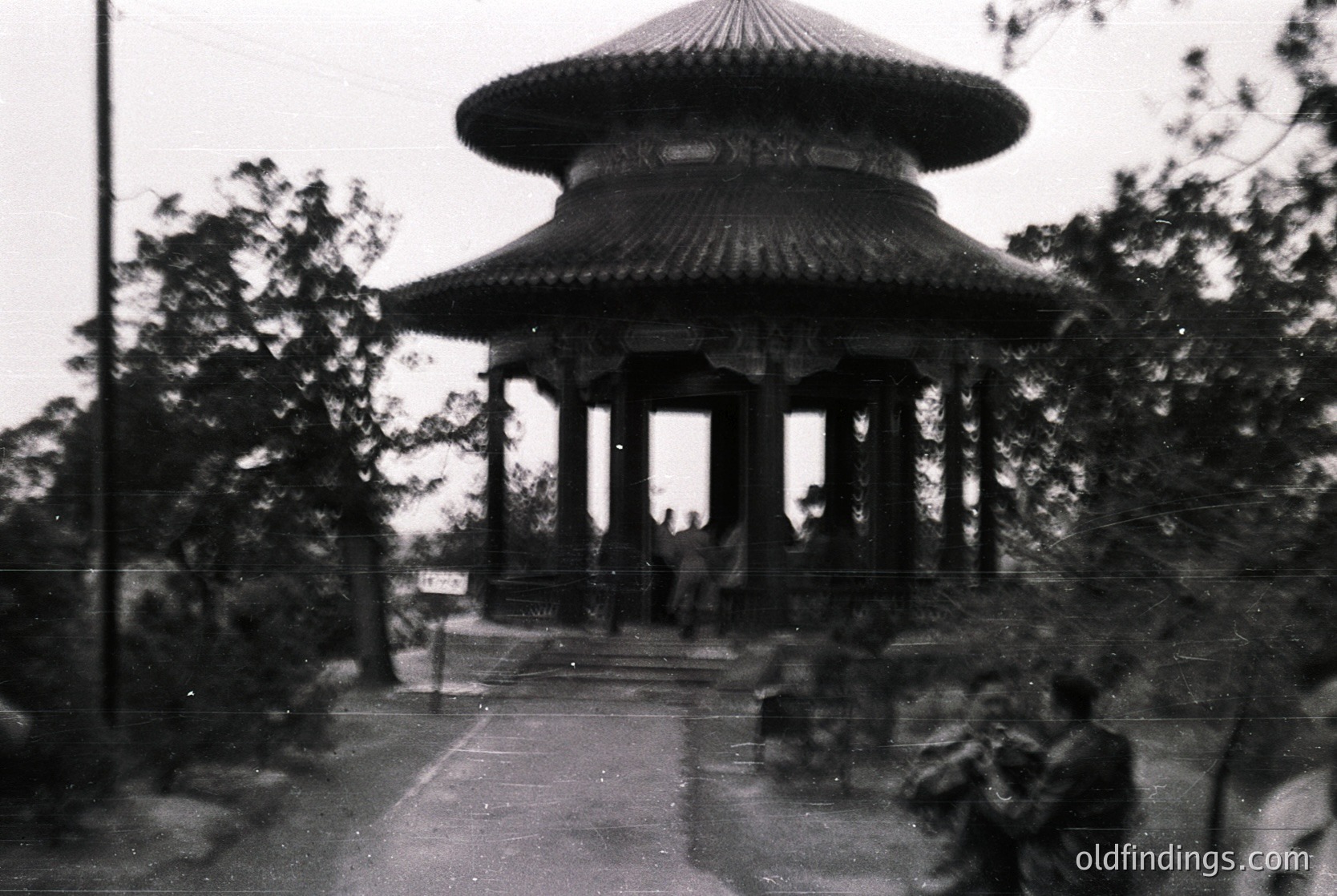 Vintage black-and-white photo of a multi-tiered pavilion with curved, tiled roof and ornate wooden columns, likely East Asian-inspired. Pathway lined with trees and benches, with blurred figures suggesting a park or public space. Mid-20th century architectural style ().