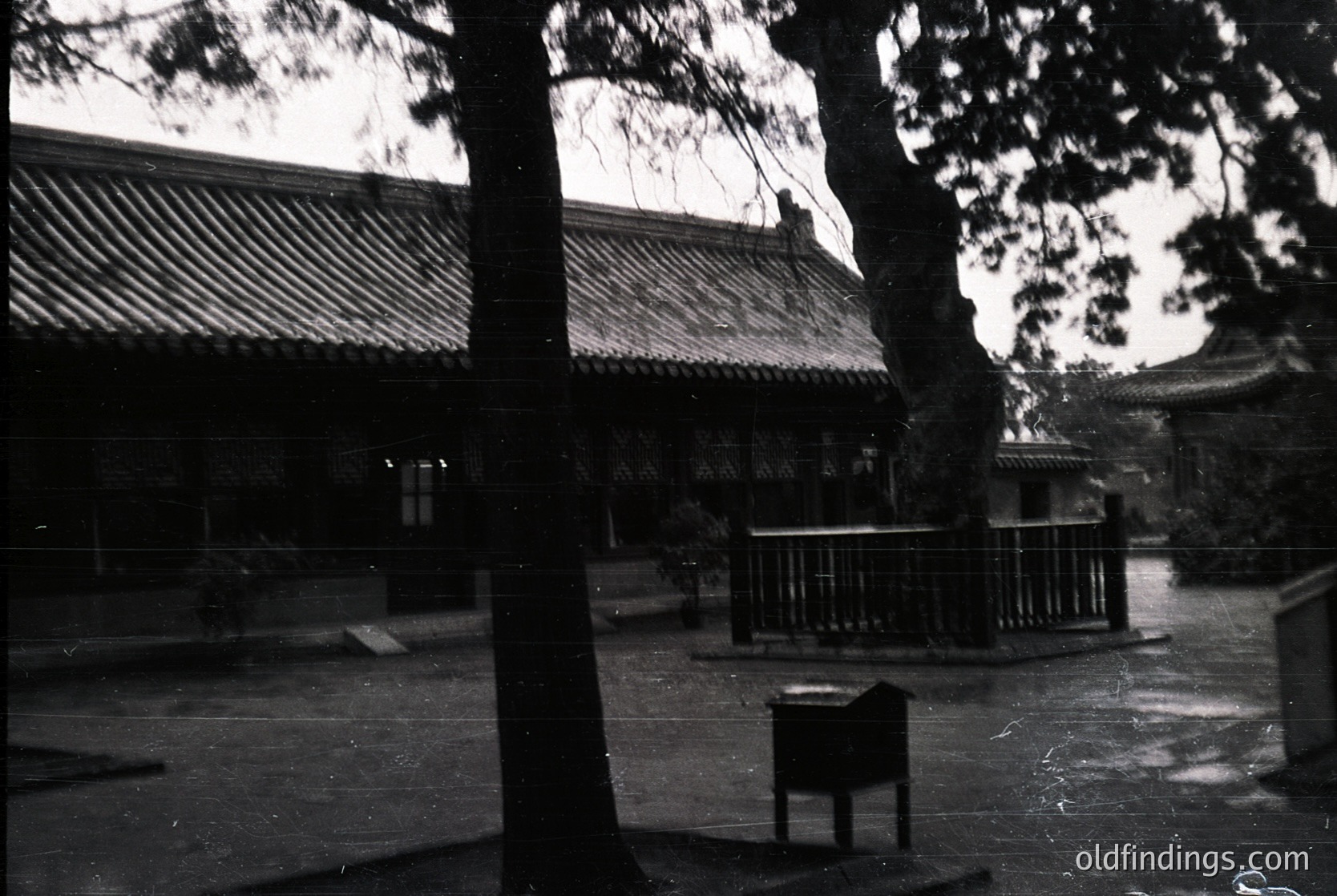 Black-and-white courtyard featuring traditional East Asian architecture with tiled roofs and wooden beams. Open pavilion with lattice railings and a small signpost in the center. Dense tree framing the left side, suggesting a serene, enclosed space. Likely a temple or historical site,