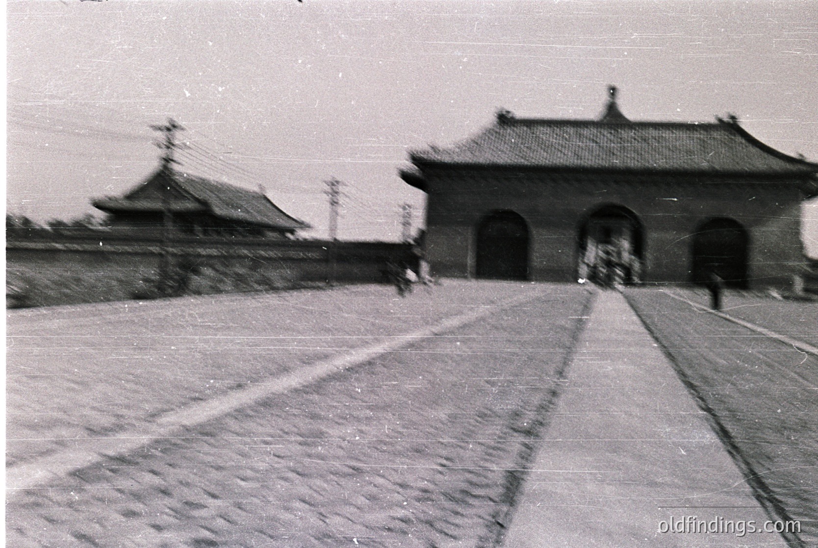 Vintage black-and-white photo of traditional East Asian gate architecture with curved, tiled roofs and arched entryways. Pedestrians walk along a wide, paved pathway flanked by low stone walls. Likely mid-20th century, possibly or .