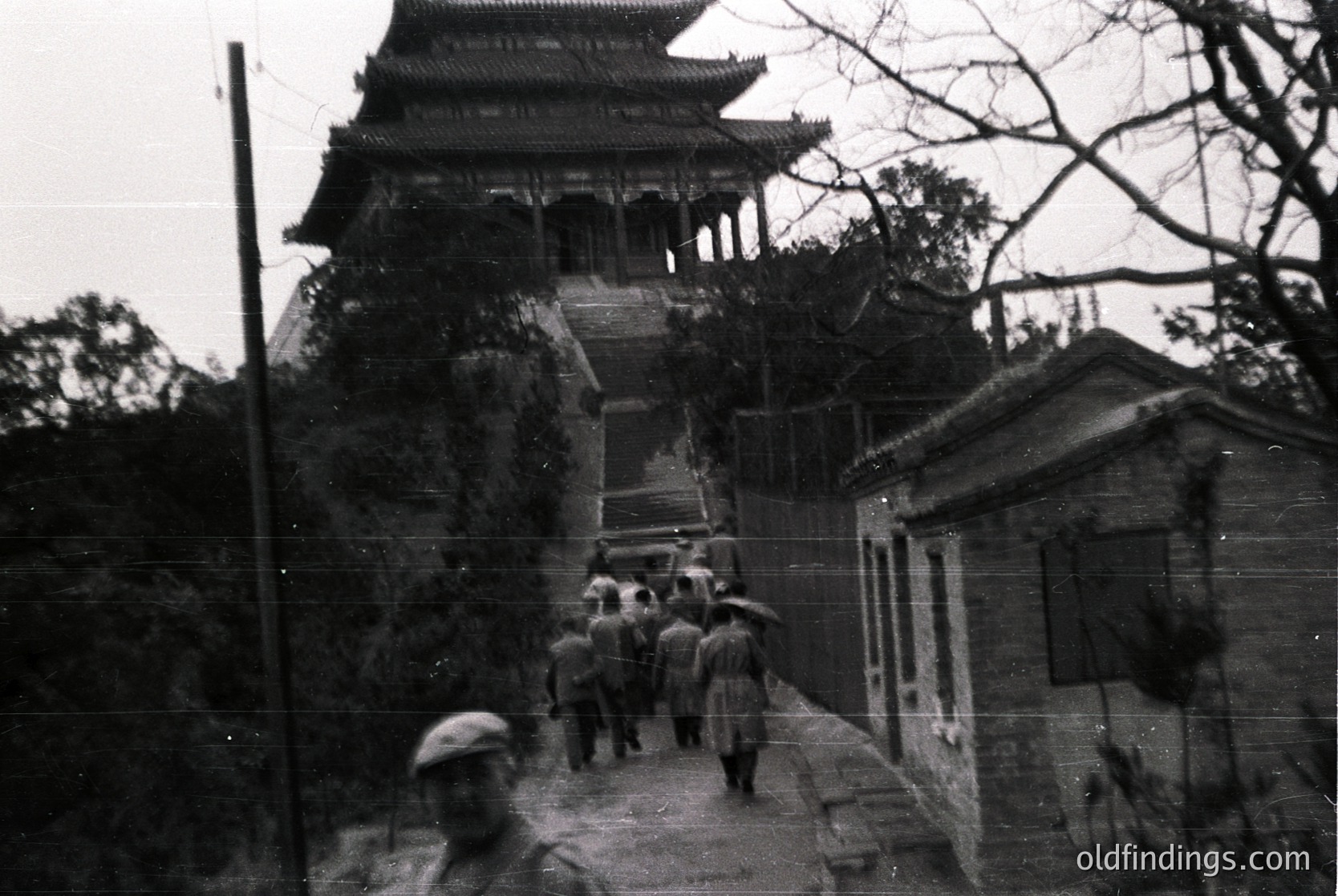 Vintage black-and-white photo of a traditional East Asian pagoda-style tower with tiered, curved eaves. A group of people in 1950s-era clothing ascends a stone pathway flanked by dense foliage. Blurred foreground figure in cap suggests candid, documentary-style capture.