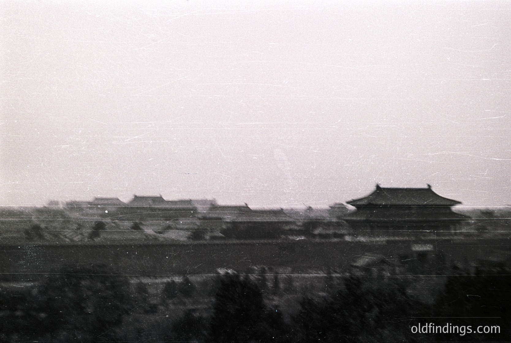 Vintage black-and-white photo of traditional East Asian palace architecture with tiled roofs and curved eaves, likely from the Ming or Qing dynasty (14th–20th century). Symmetrical pavilions line a fortified wall, framed by dense foliage below.
