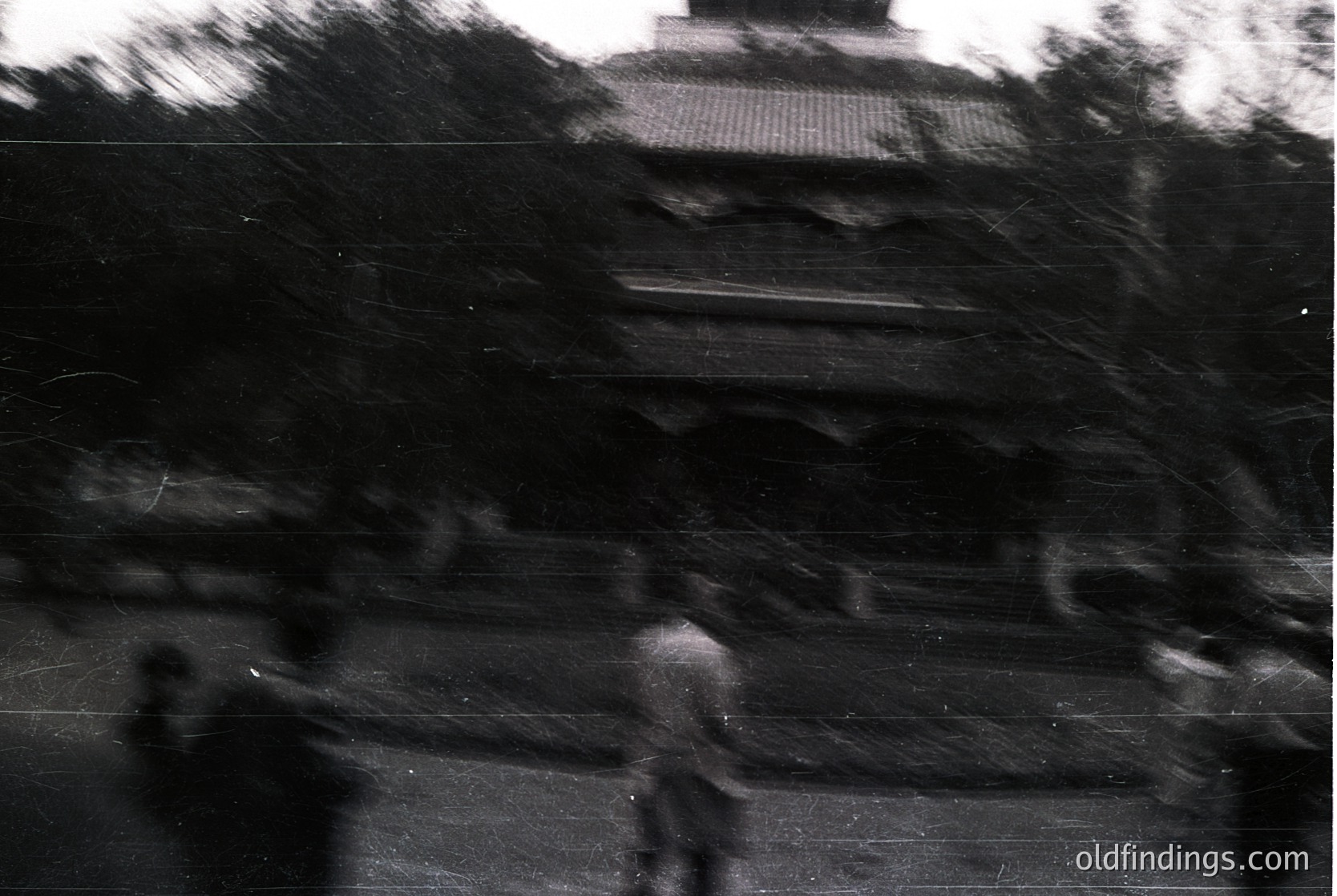 Vintage black-and-white shot of a blurred urban street scene, likely mid-20th century. Silhouetted figures walk along a paved road flanked by indistinct buildings and trees. Architectural details suggest a European cityscape, possibly or .