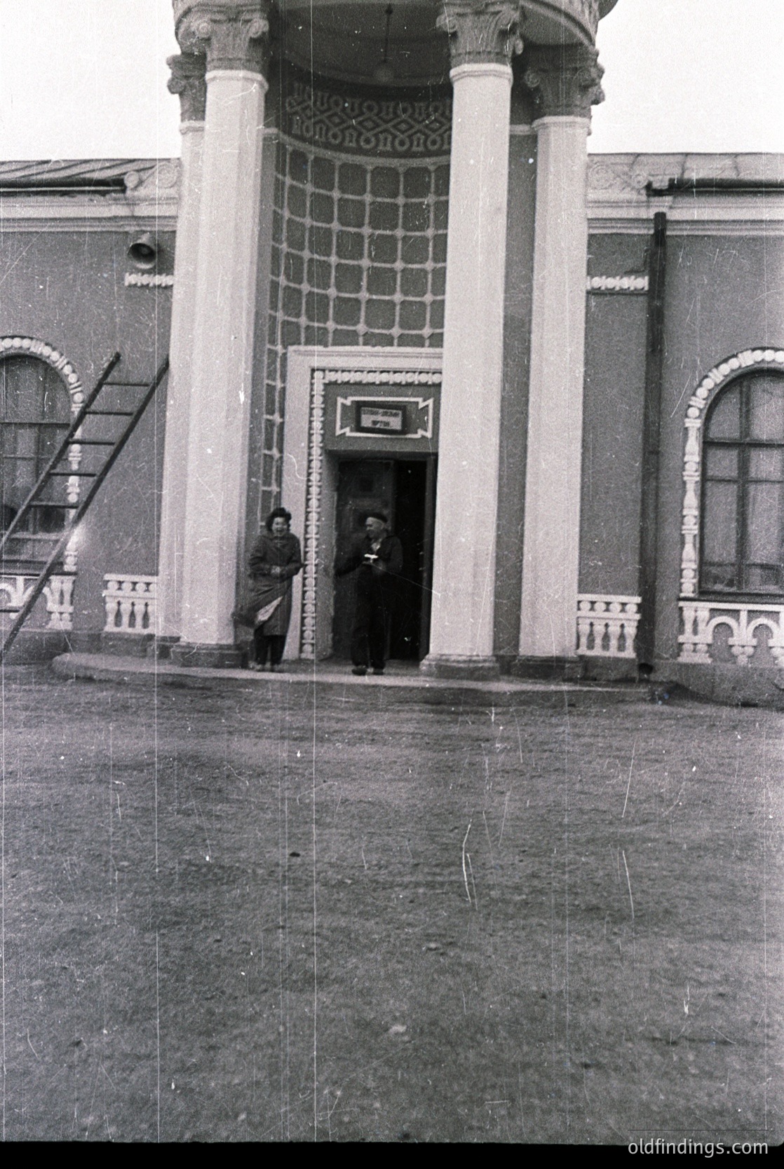 Neoclassical building entrance with two men in early 20th-century attire, standing under a portico supported by fluted columns. Ornate wrought-iron balcony railing and tiled mosaic panel above the door. Signage suggests a public or institutional function.