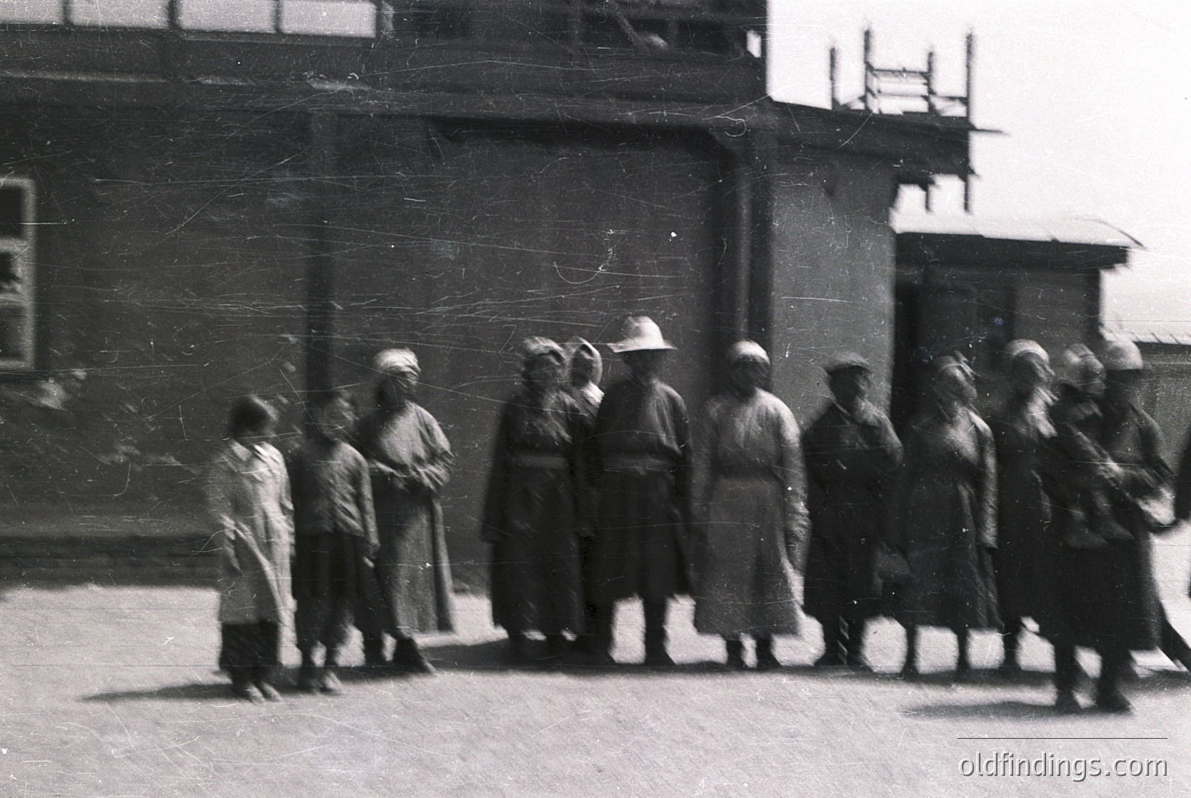 Group of women in early 20th-century attire, likely Eastern European, standing outside a brick building with scaffolding. Heavy coats, headscarves, and some wearing hats suggest cold weather. Urban setting with visible industrial elements.