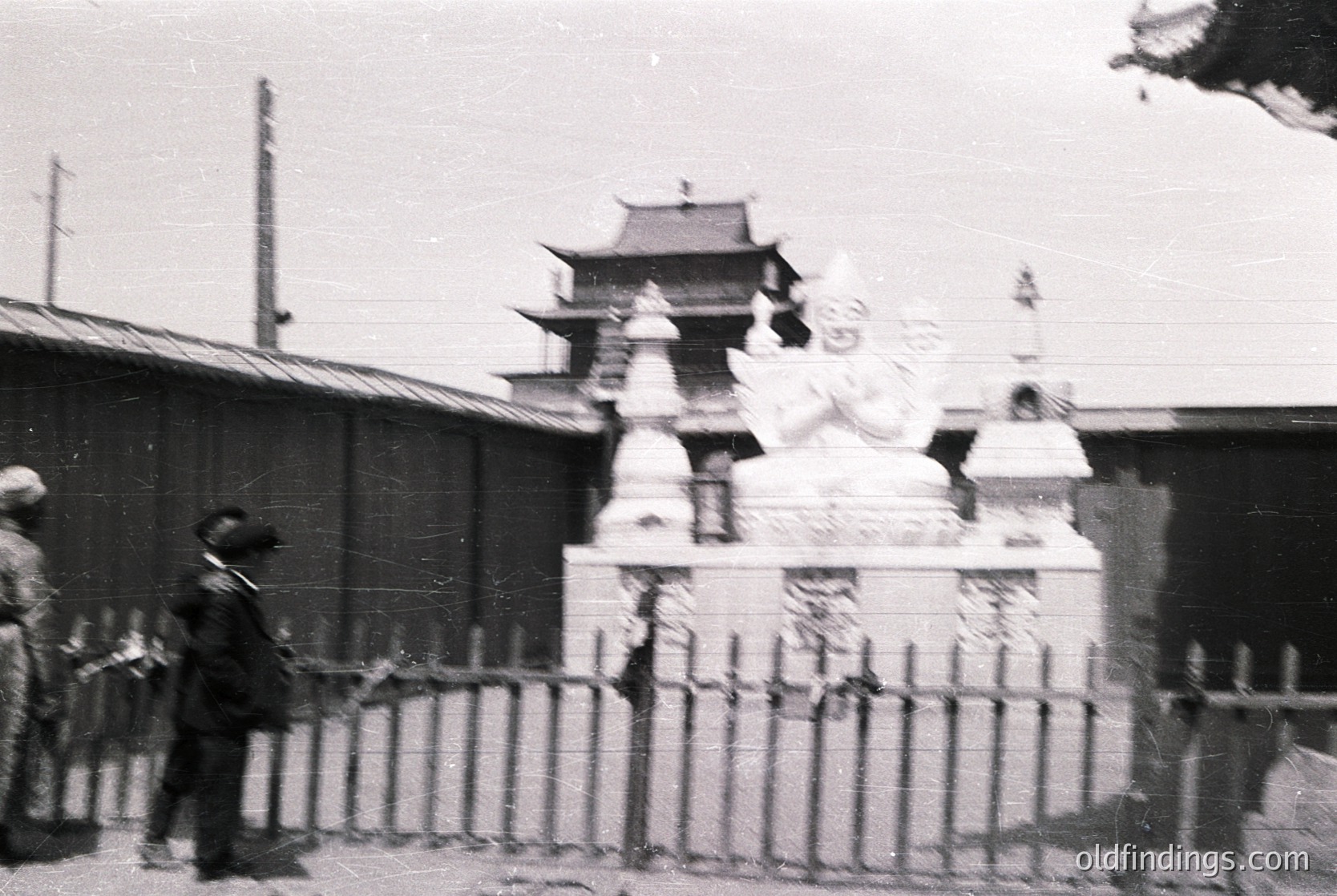 Vintage black-and-white photo of a traditional East Asian architectural gate with tiled pagoda roof, flanked by stone lion statues. A man in 1950s-era suit walks near a metal fence. Urban setting with partial buildings visible.