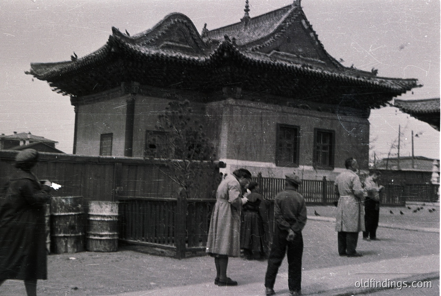 Black-and-white street scene featuring a traditional East Asian-style building with upturned eaves and ornate roof details, likely from the early-to-mid 20th century. Group of people in vintage clothing—men in coats, women in long dresses—standing near a wrought-iron fence. Urban setting with visible street and industrial barrels.