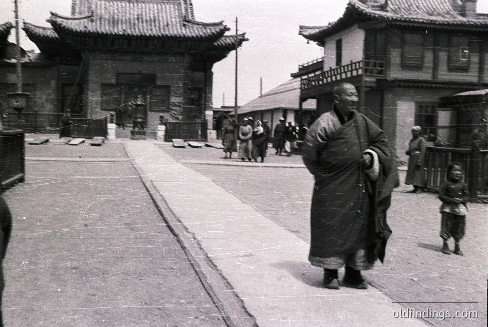 Monk in traditional robe strides along paved street flanked by traditional East Asian architecture with tiled roofs and wooden beams. Mid-20th century urban scene, likely Japan or Korea.
