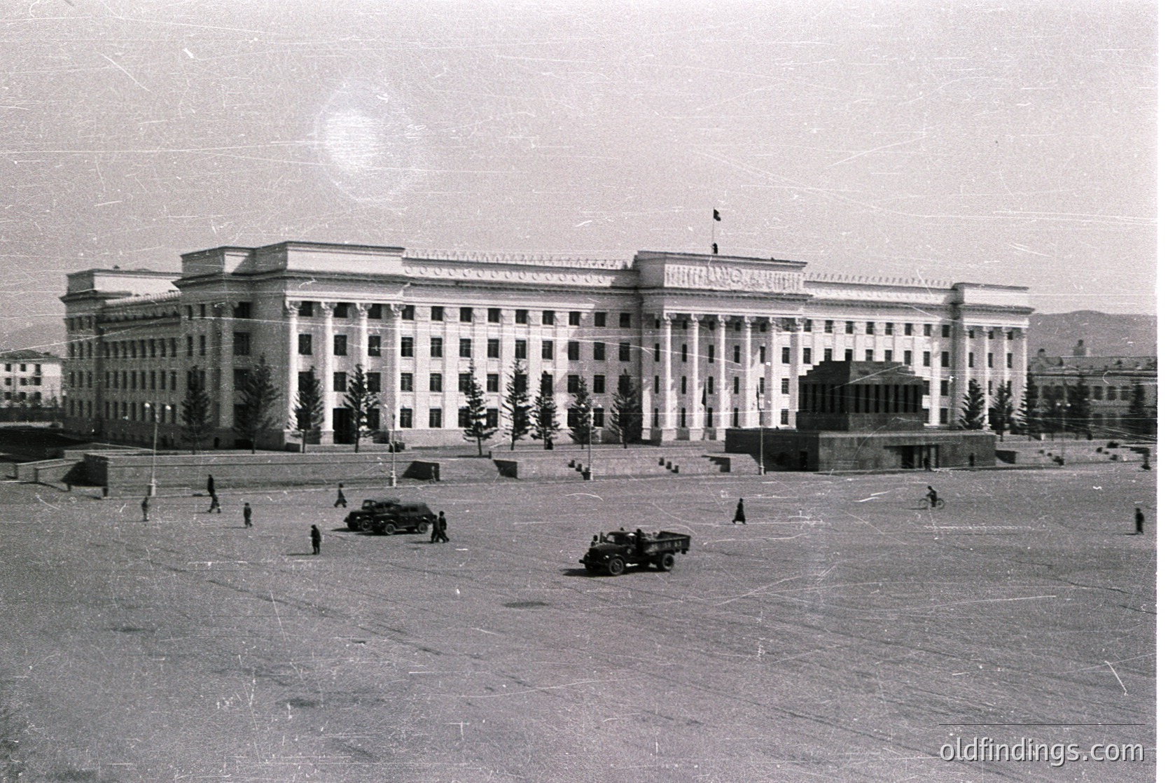 Neoclassical-style Soviet-era building with symmetrical columns and a central entrance, likely a government or institutional structure. Open plaza with sparse pedestrians and a military vehicle in foreground. --- *Note: The building’s design aligns with mid-20th-century Soviet architectural trends, and the military vehicle suggests a Cold War context. The setting resembles Sofia’s central plaza, though further verification would confirm location.*