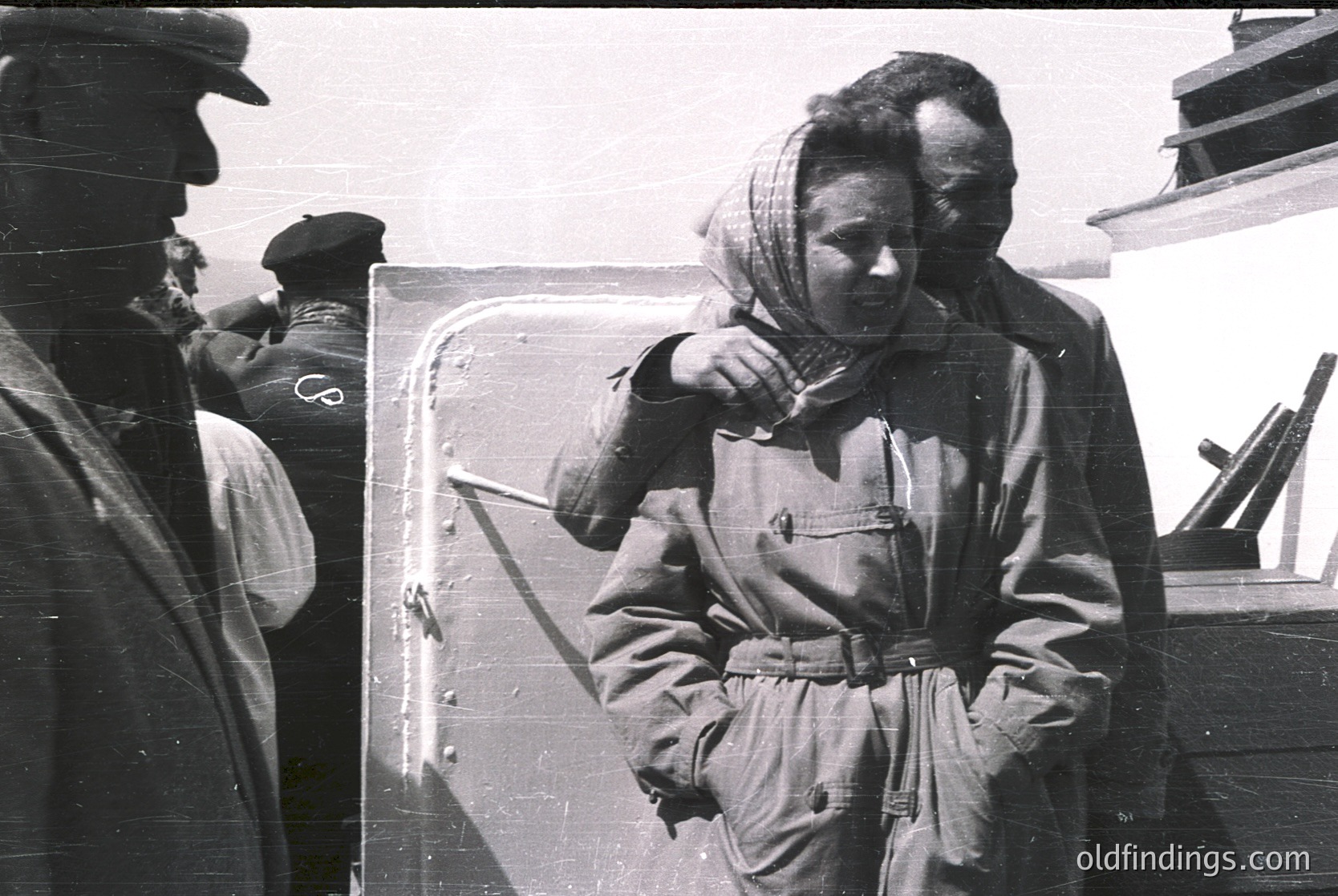 Black-and-white photo of three individuals on a ship’s deck, mid-20th century. A woman in a heavy coat and headscarf leans on a metal hatch, while a man in a peaked cap points toward her. Another man in uniform stands partially obscured. Maritime setting suggests wartime or post-war era.