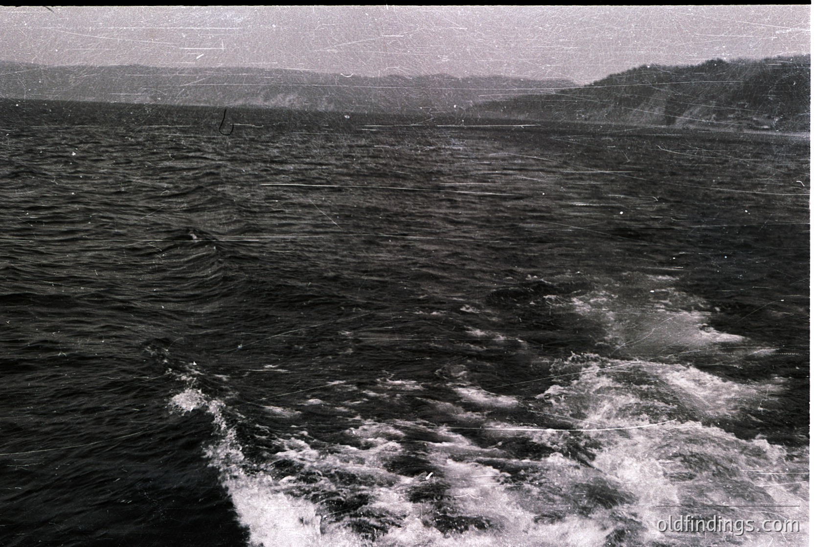 Black-and-white maritime scene showing turbulent waters with visible wake from a vessel. Distant shoreline with forested hills under overcast skies. Vintage monochrome aesthetic suggests mid-20th century. Ideal for historical maritime or coastal research.
