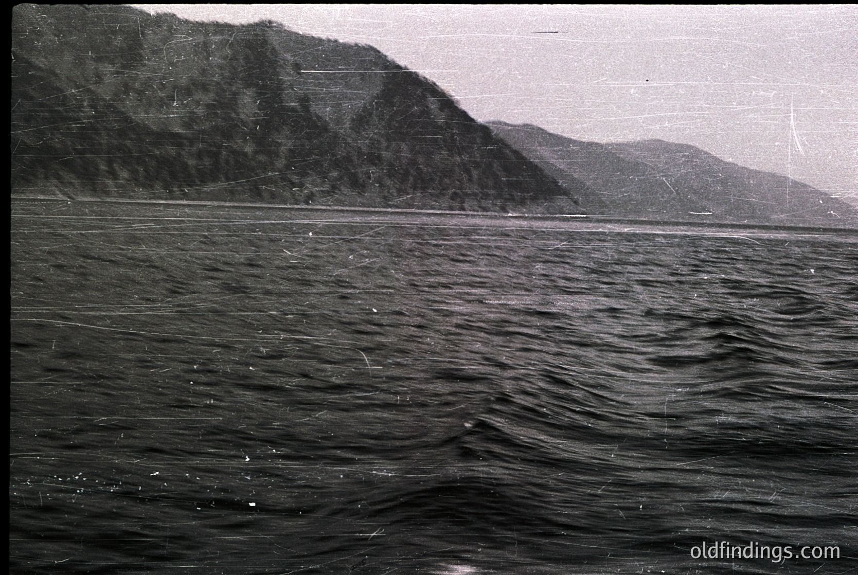 Black-and-white coastal scene featuring rugged cliffs meeting choppy waters. Dense forest covers the steep terrain, with minimal visible human presence. Likely mid-20th century due to grainy texture and lighting. Ideal for vintage travel or nature stock imagery.
