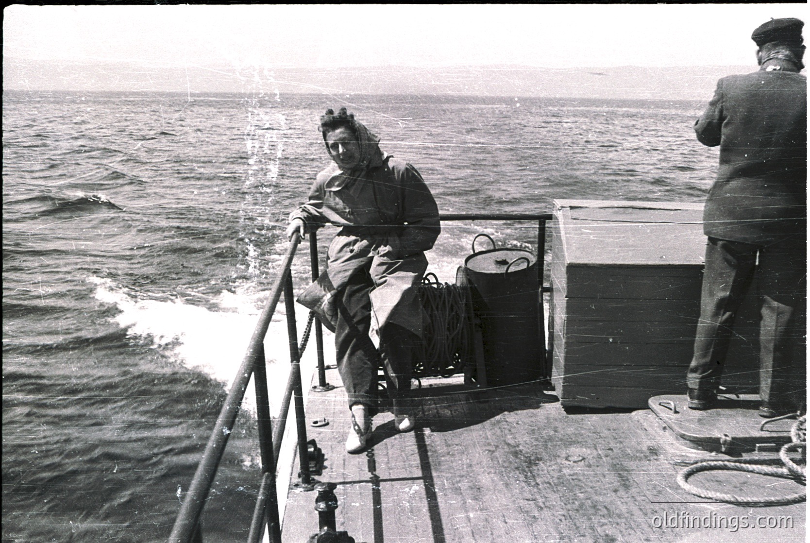 Black-and-white maritime scene: two individuals on a ship’s deck, one seated on a rail holding a bag, the other standing beside a wooden box. Waves and open sea in background. Mid-20th century naval or merchant vessel.