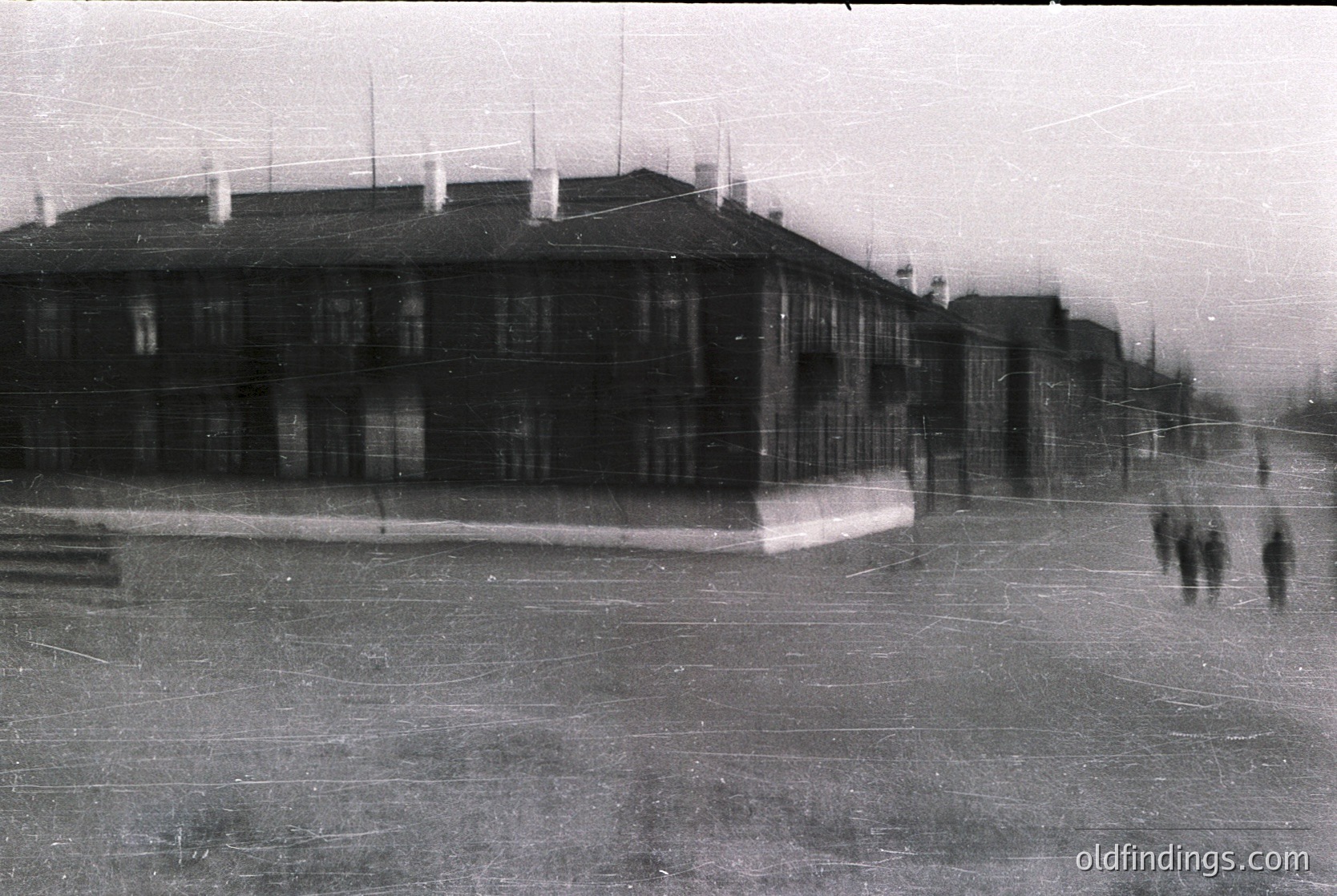 Vintage black-and-white shot of a Soviet-era institutional building with Soviet-style architecture—multi-story, symmetrical, and utilitarian. Blurred figures suggest a public square or courtyard. Likely Eastern Bloc, mid-20th century.