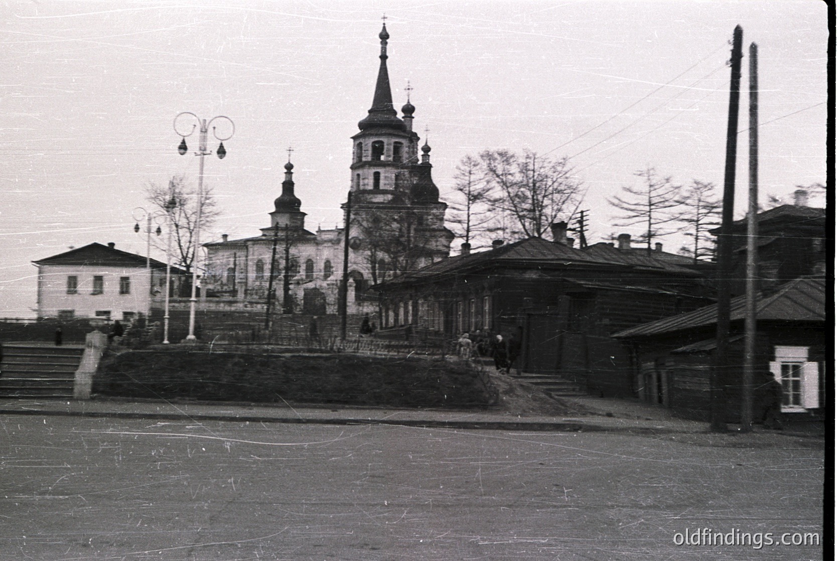 Black-and-white street view of a Russian Orthodox church with twin spires and a central bell tower, featuring intricate detailing. Surrounding buildings exhibit Soviet-era architecture, with a mix of residential and utilitarian structures. Pedestrians in 1950s–60s attire walk along a paved road.