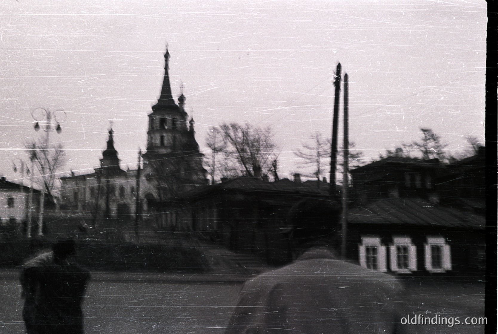 Vintage black-and-white photo of a Soviet-era Orthodox church with a tall, pointed steeple and smaller bell towers, framed by bare winter trees. Foreground shows indistinct figures in heavy coats, likely 1950s–1970s Soviet Union.
