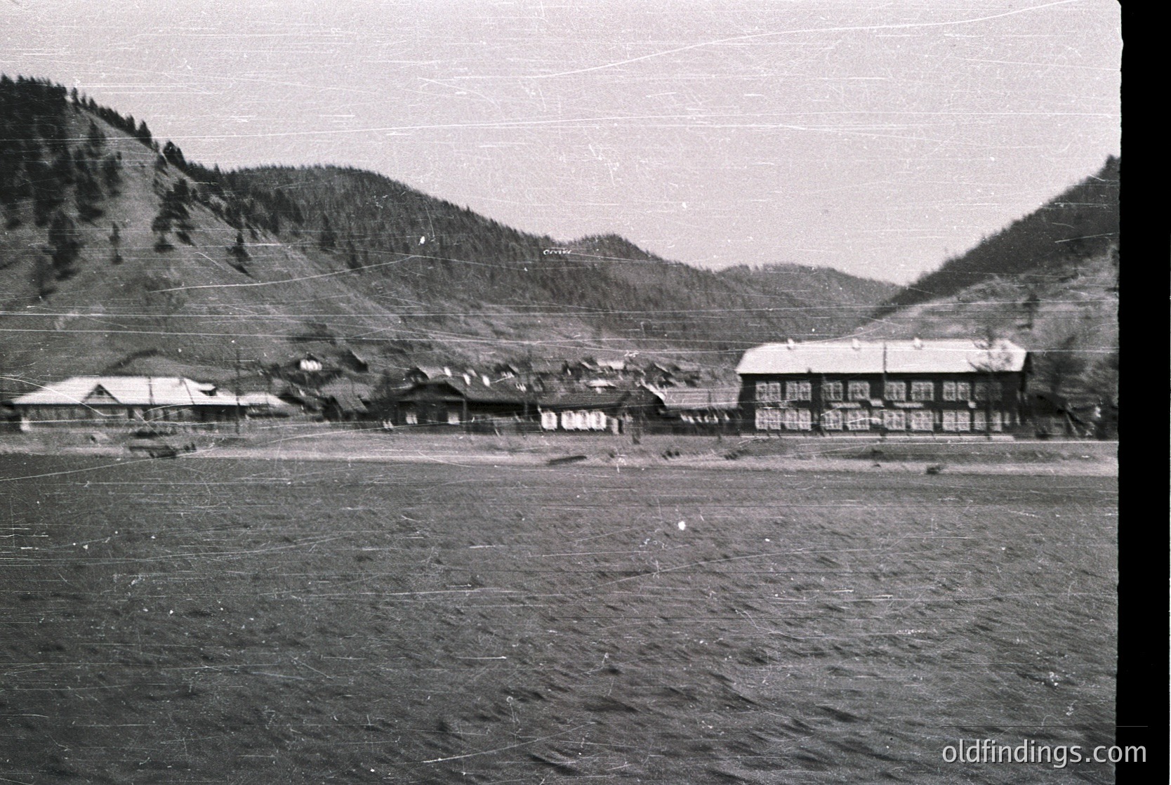 Mid-20th century alpine village nestled between forested slopes. Wooden chalets with gabled roofs line a flat valley floor, surrounded by dense pine forests. Likely European, possibly or , .