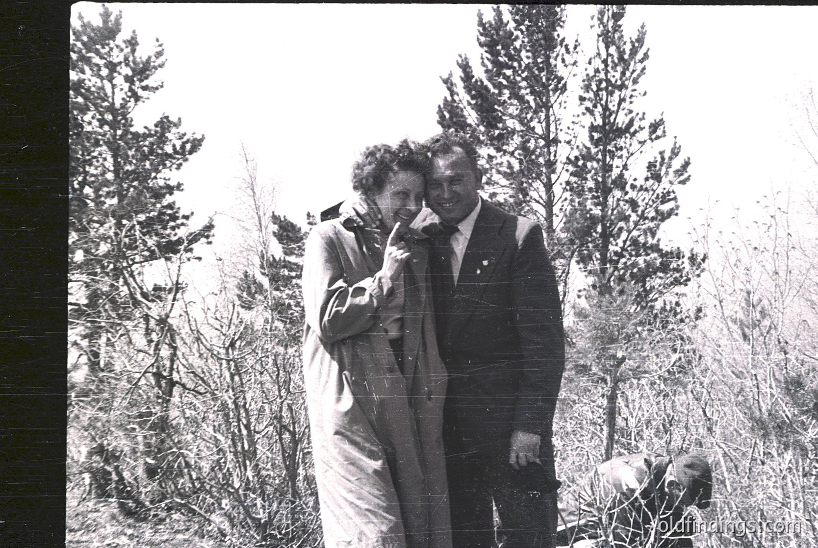 Two individuals in mid-20th century attire pose outdoors in a forested area, likely mid-1950s–1960s. Man wears a suit with a tie, woman a long coat and headscarf. Blurred background suggests motion or low-resolution film.